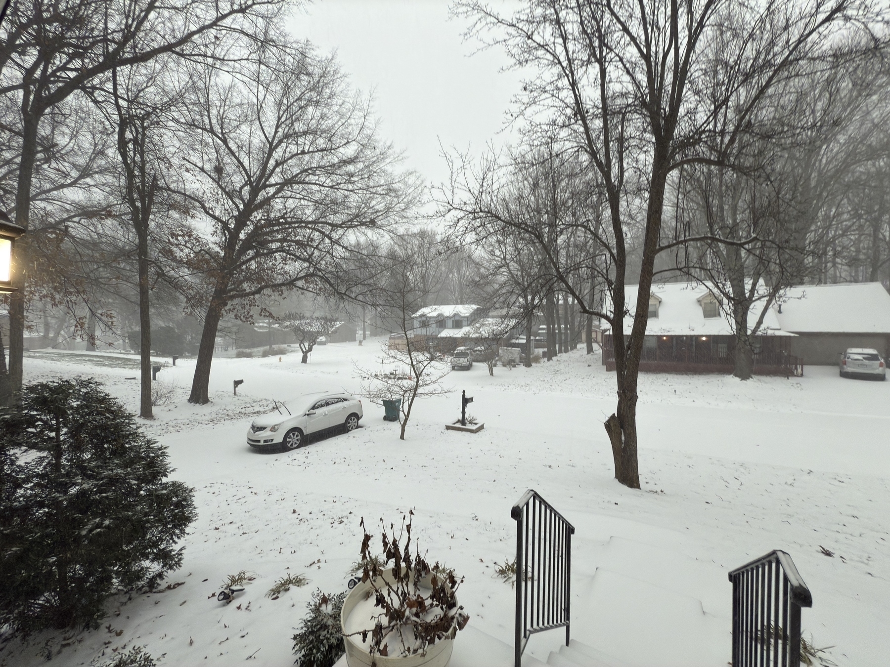 A snowy suburban scene features a car parked on a street with houses surrounded by bare trees.