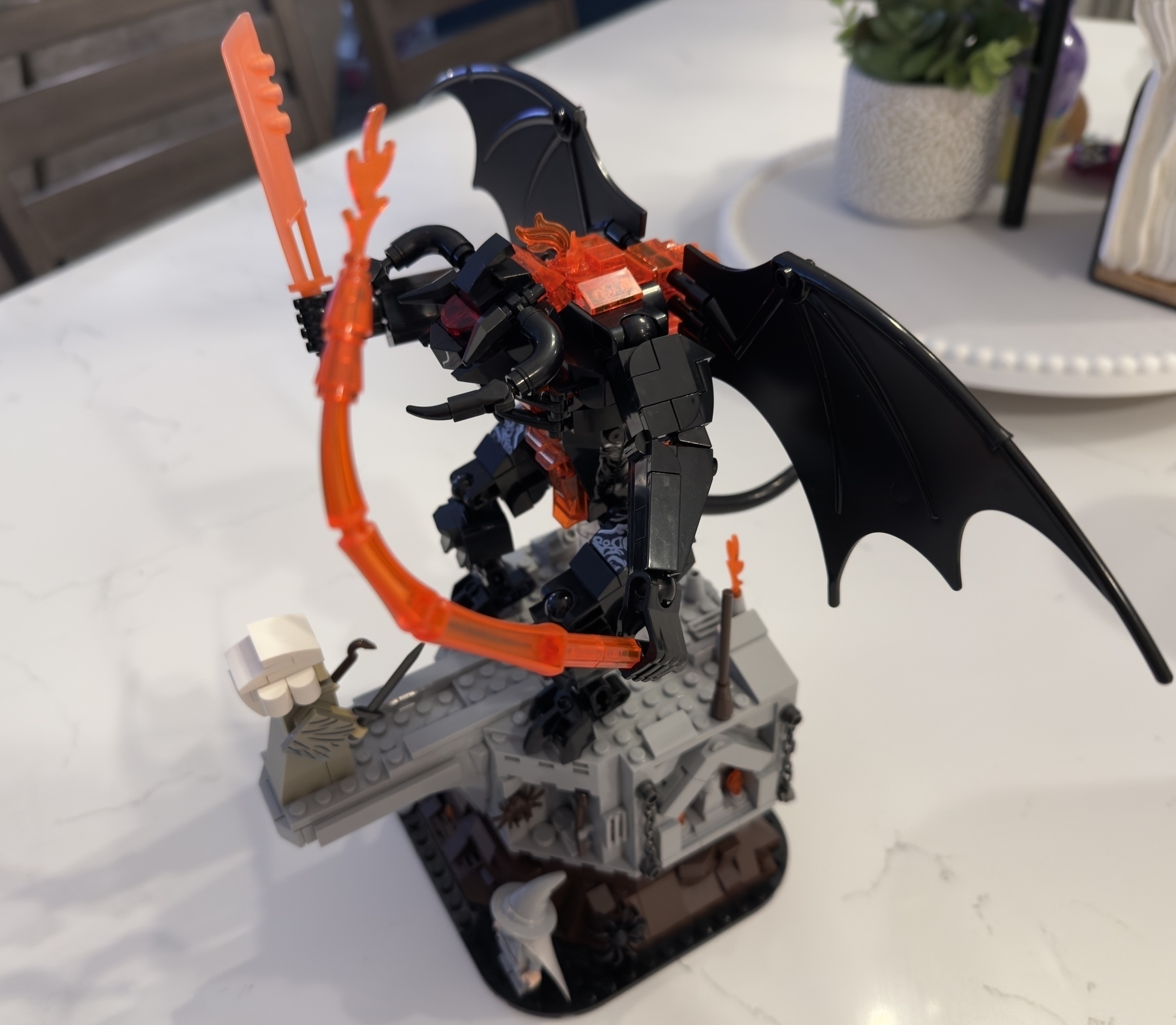 A LEGO build depicts a winged creature with fiery weapons on a rocky base, poised near a small robed figure.