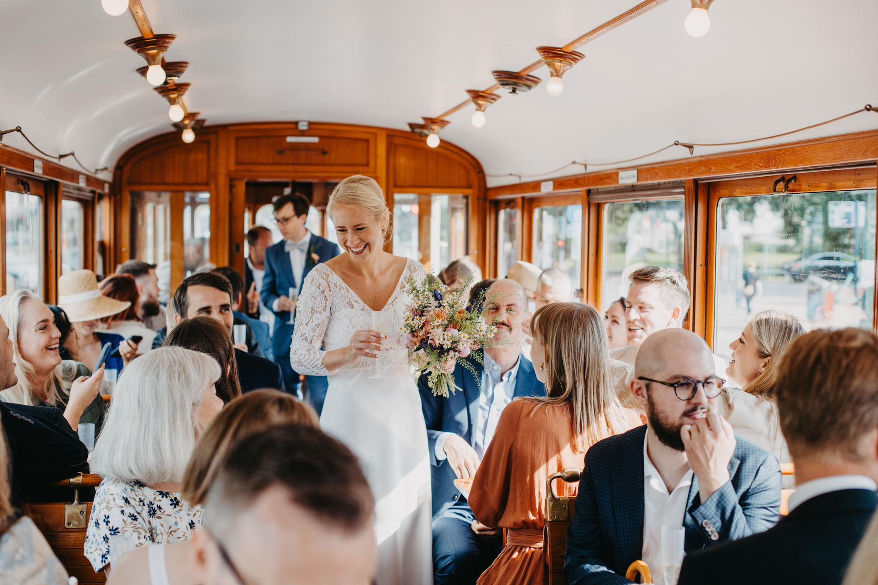 Auto-generated description: A bride in a white dress is holding a bouquet and standing in a tram filled with smiling, well-dressed guests.