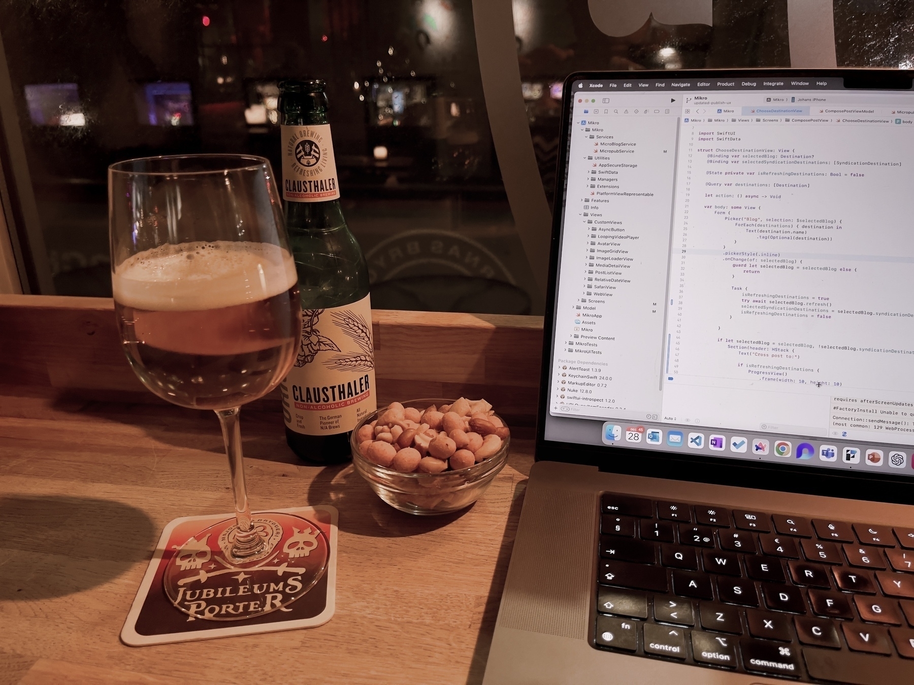 A wooden table with a glass of beer on a coaster, a bottle, a bowl of nuts, and an open laptop displaying code.