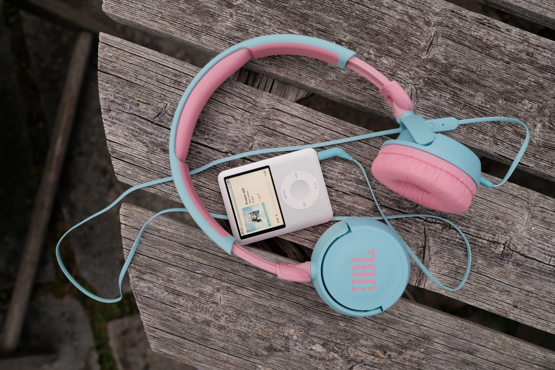 A pastel blue and pink pair of JBL headphones is resting on a weathered wooden surface alongside a white iPod displaying album art for Frozen. 