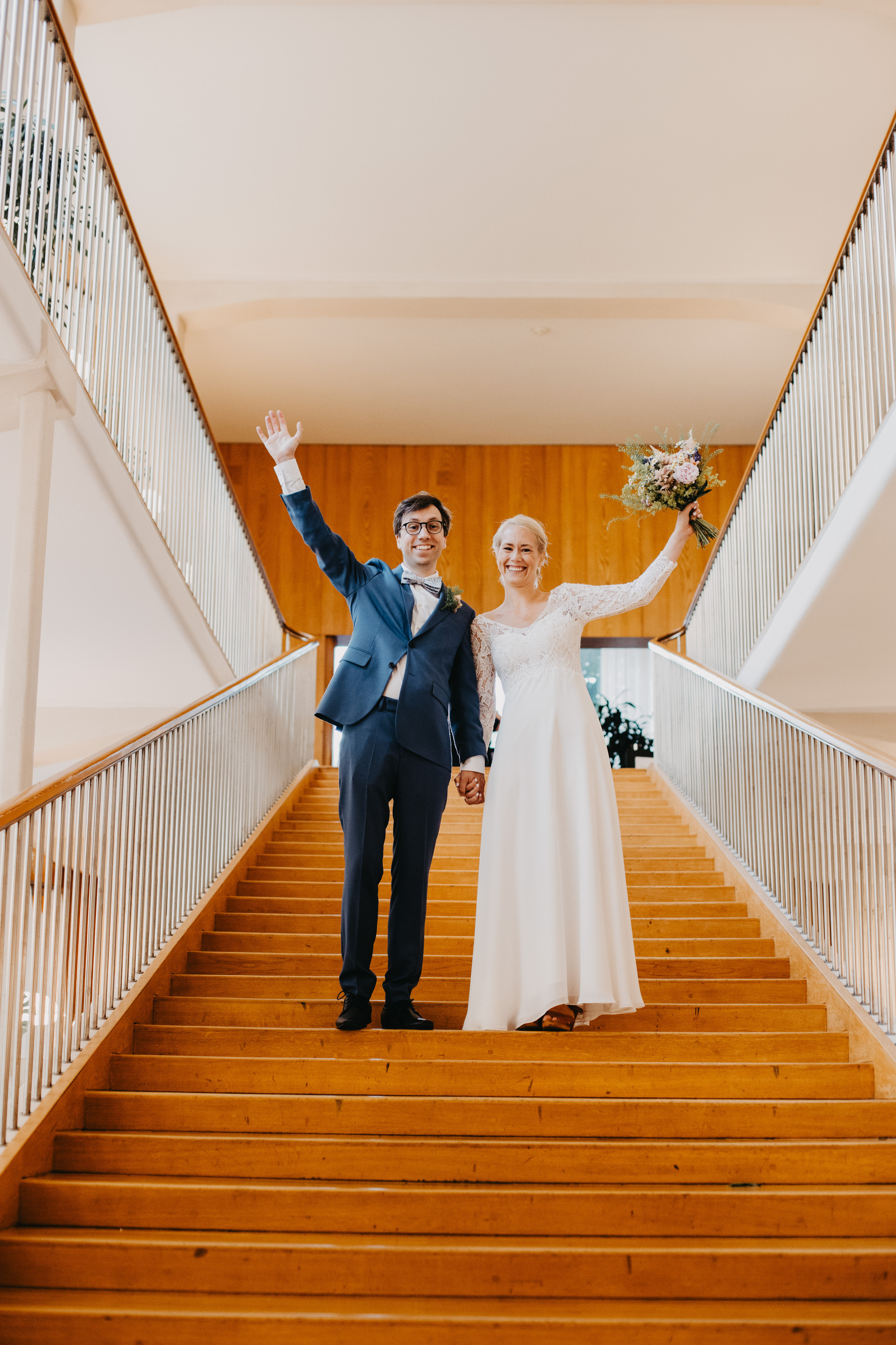Auto-generated description: A newlywed couple is standing at the top of a staircase, smiling and waving, with the bride holding a bouquet.