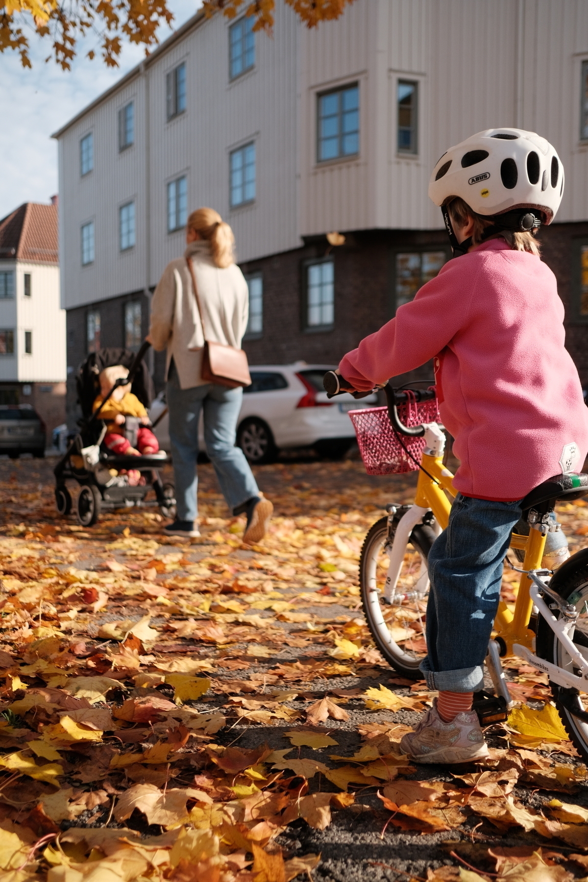 Barn på cykel i förgrunden med höstlöv på marken och en mamma med barnvagn i bakgrunden.