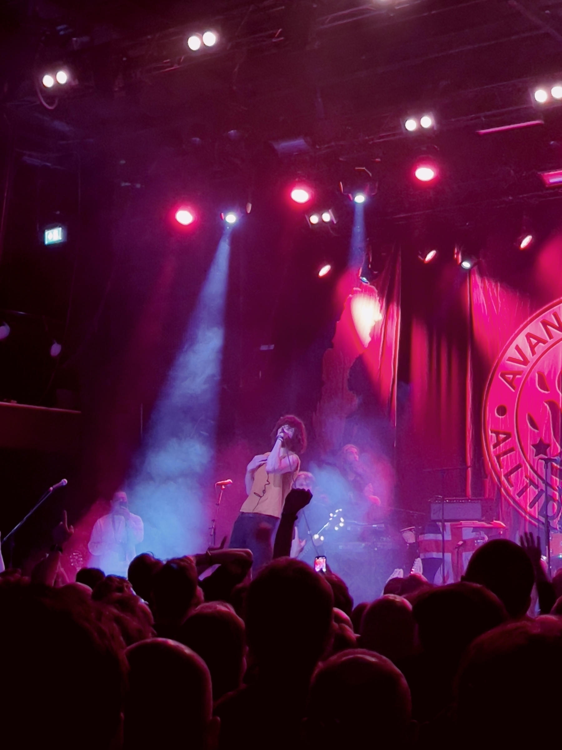 A band performs on a dimly lit stage with vibrant purple and red lighting and a large crowd watching.