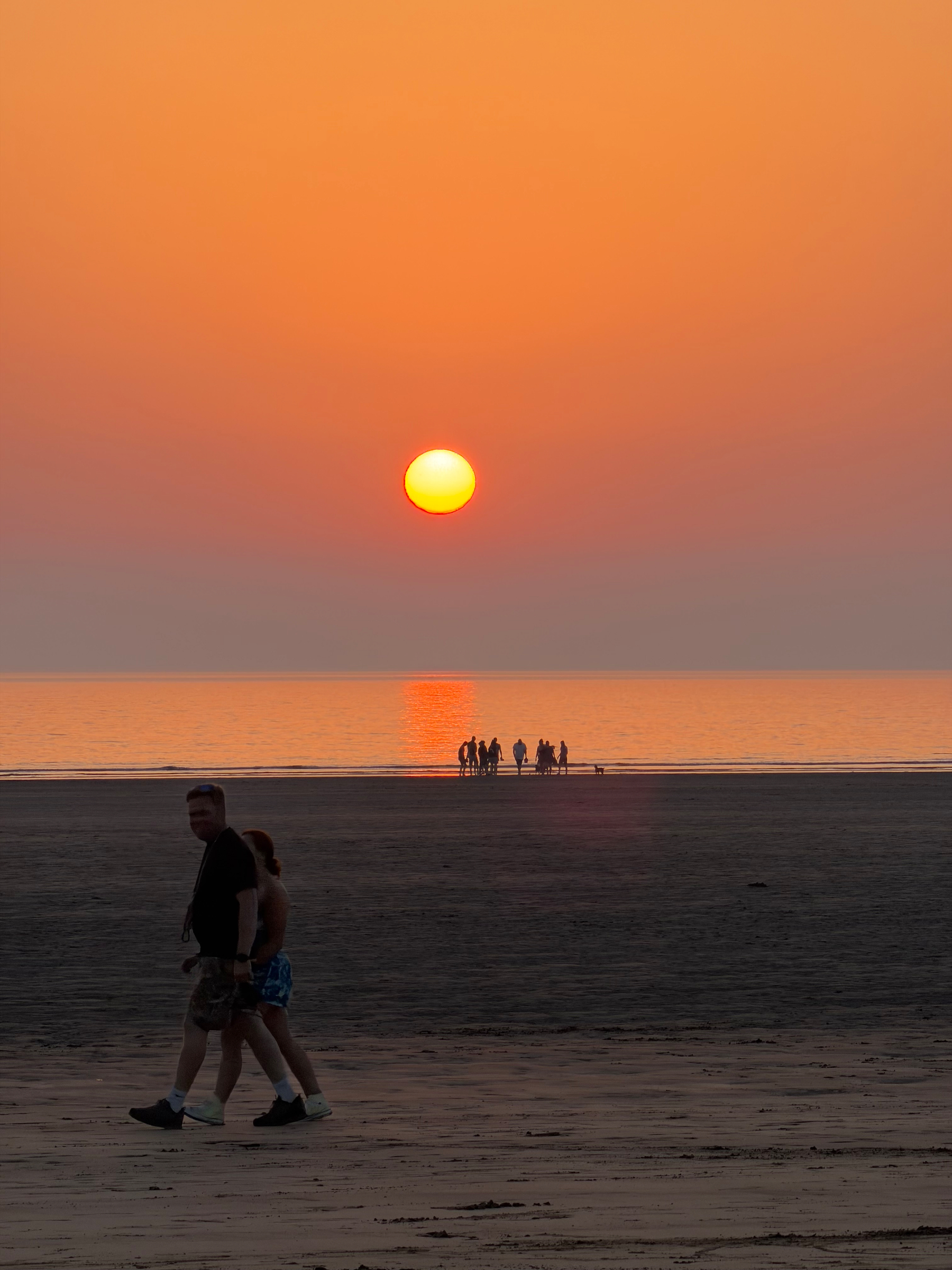 A group of people is gathered near the water's edge while two individuals walk along the beach during a vibrant orange sunset.
