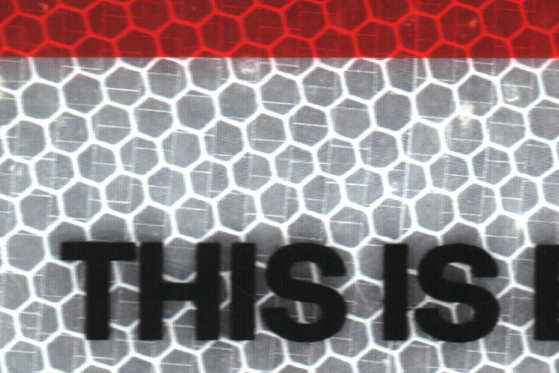 Close up of a reflective sign. Red stripe at the top, reflective hexagons and the words "this is" in caps