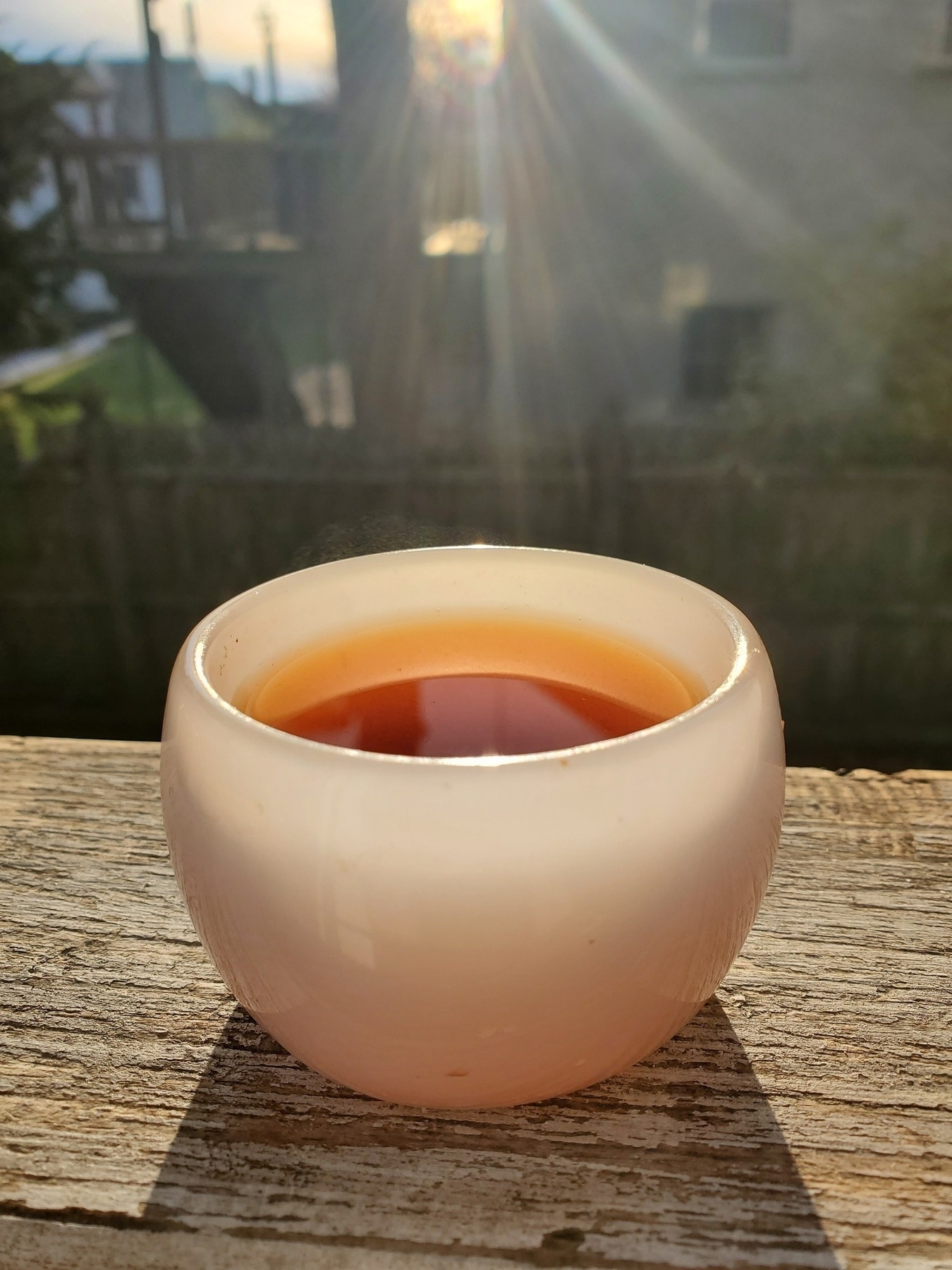 A small white porcelain cup in the chinese or japanese teacup style, full of a rich dark tea, on a porch railing, illuminated by the morning sun.