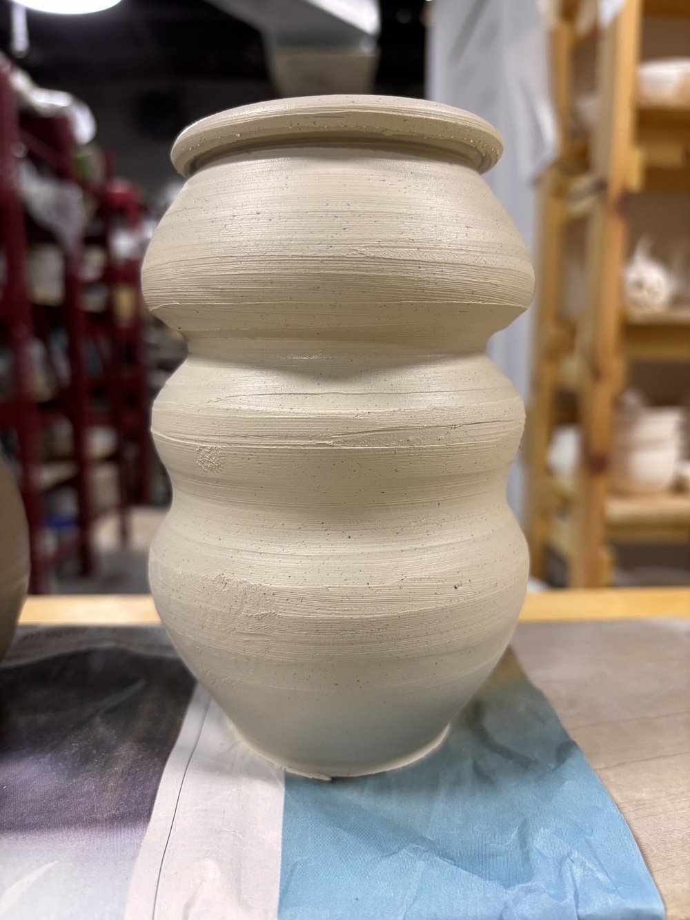 A hand-thrown clay vase with a rounded, undulating shape sits on a table in a pottery studio.