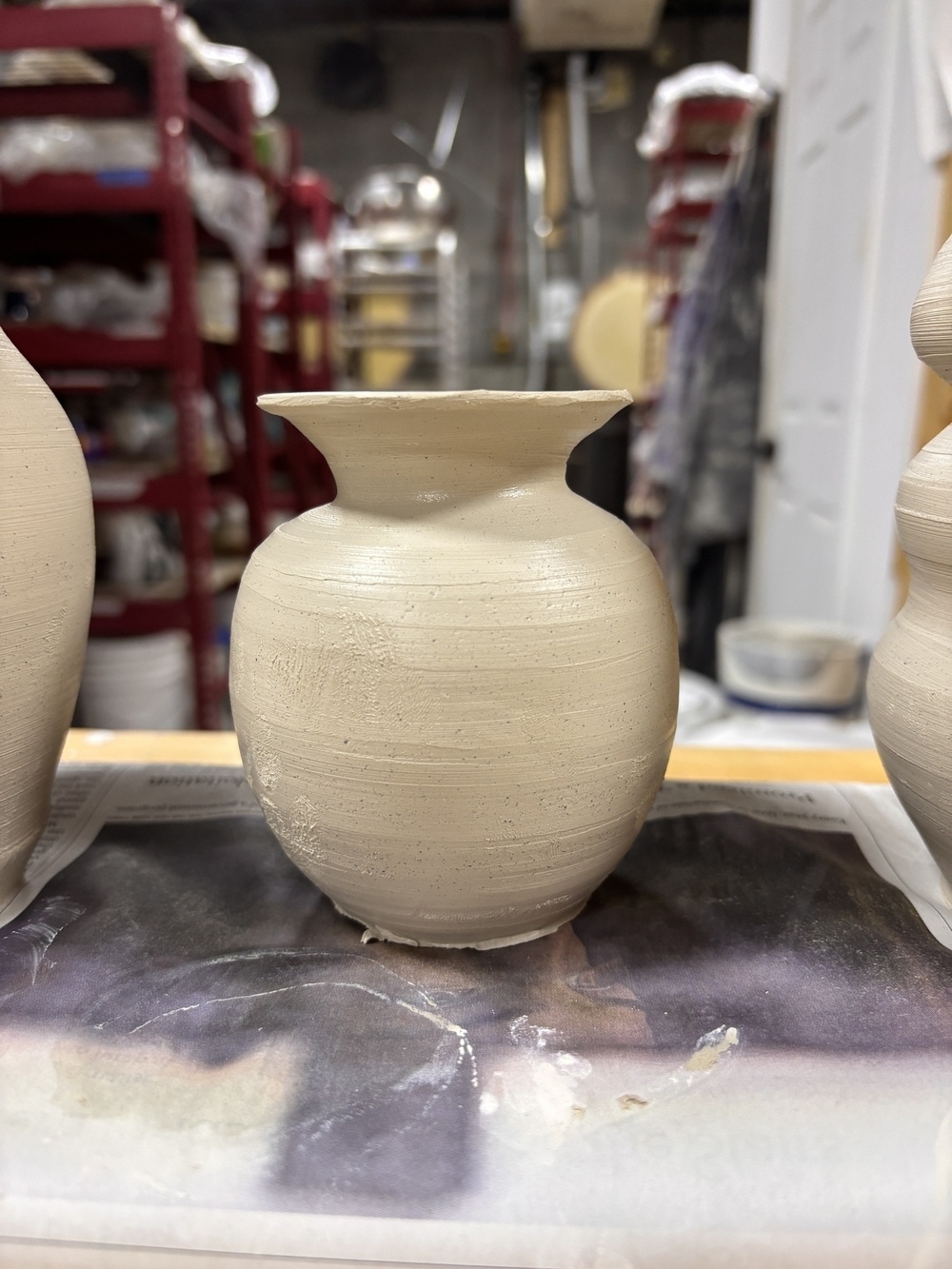 A clay vase, drying on a workbench, is surrounded by pottery tools and materials.