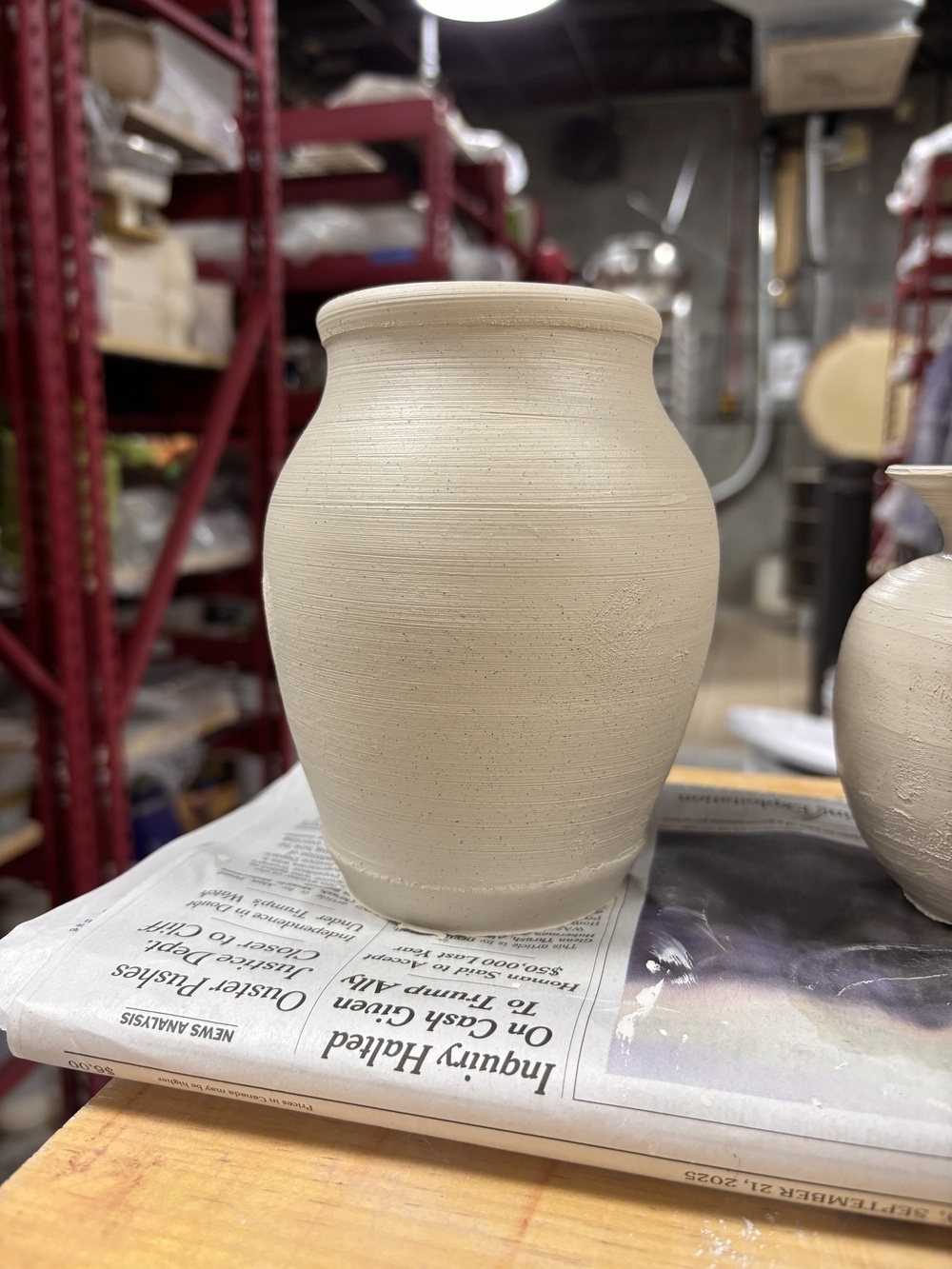 A clay pot sits on a newspaper in a pottery studio surrounded by shelves and equipment.