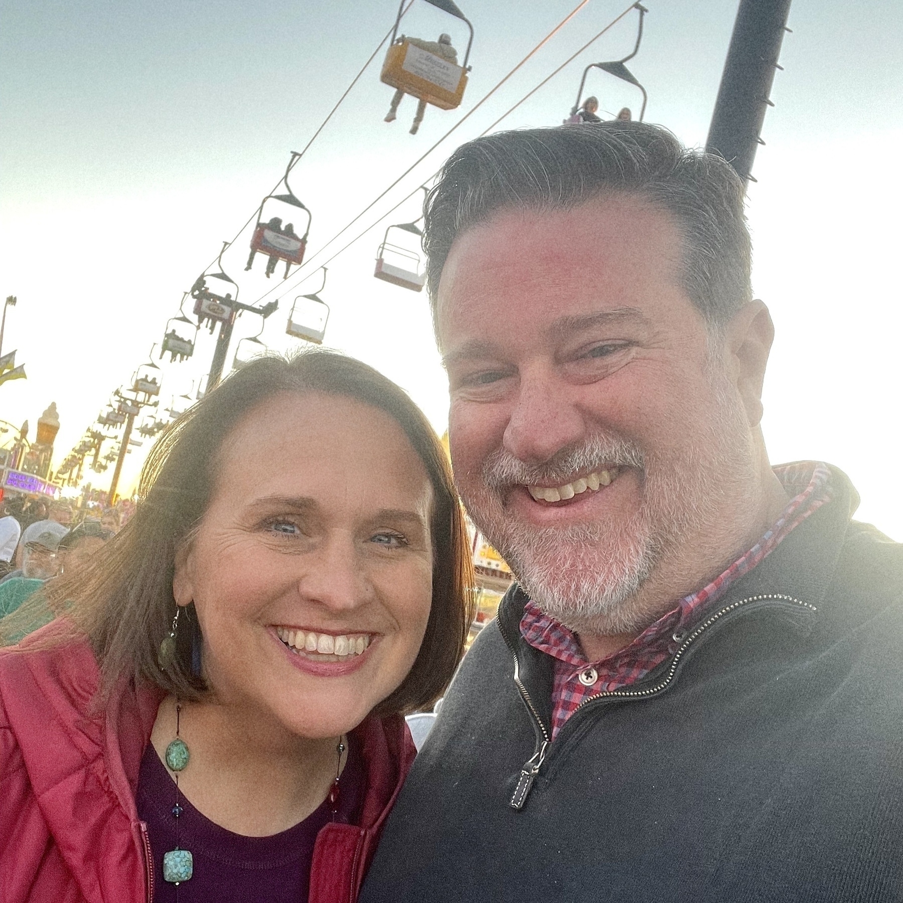 Bob and Liz take a selfie in front of a chairlift with people riding above.