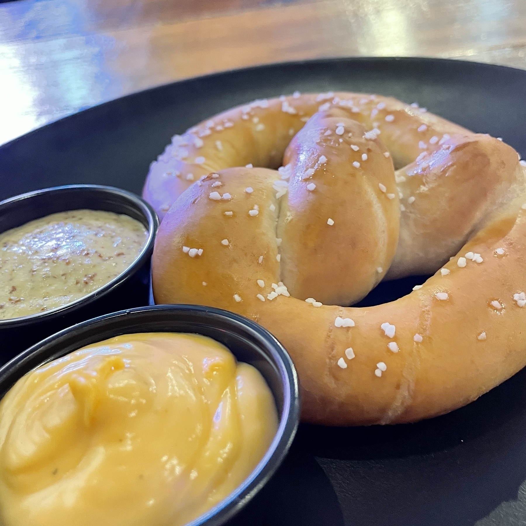 A pretzel sprinkled with salt is served on a black plate alongside two small dishes of mustard and beer cheese.