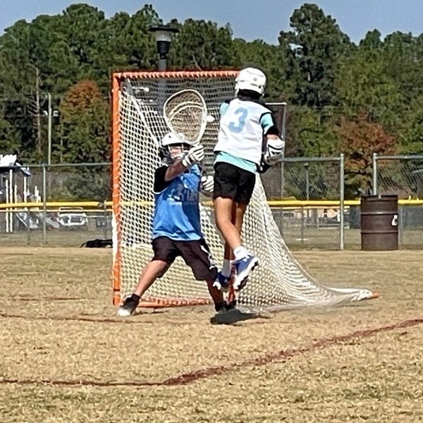 Auto-generated description: Two young athletes are engaged in a lacrosse game on a grassy field.