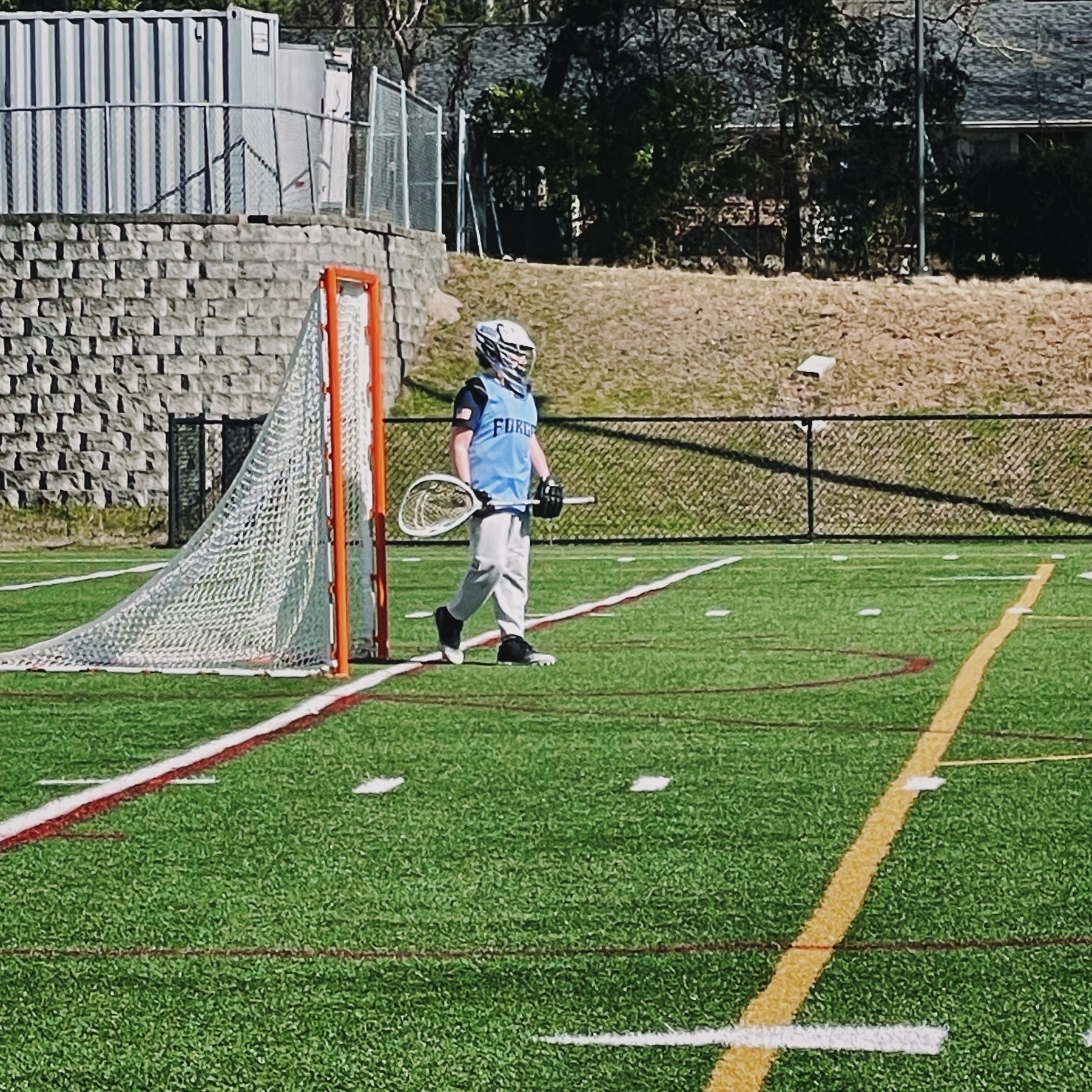 A lacrosse goalie wearing protective gear stands in front of a goal on a grassy field.