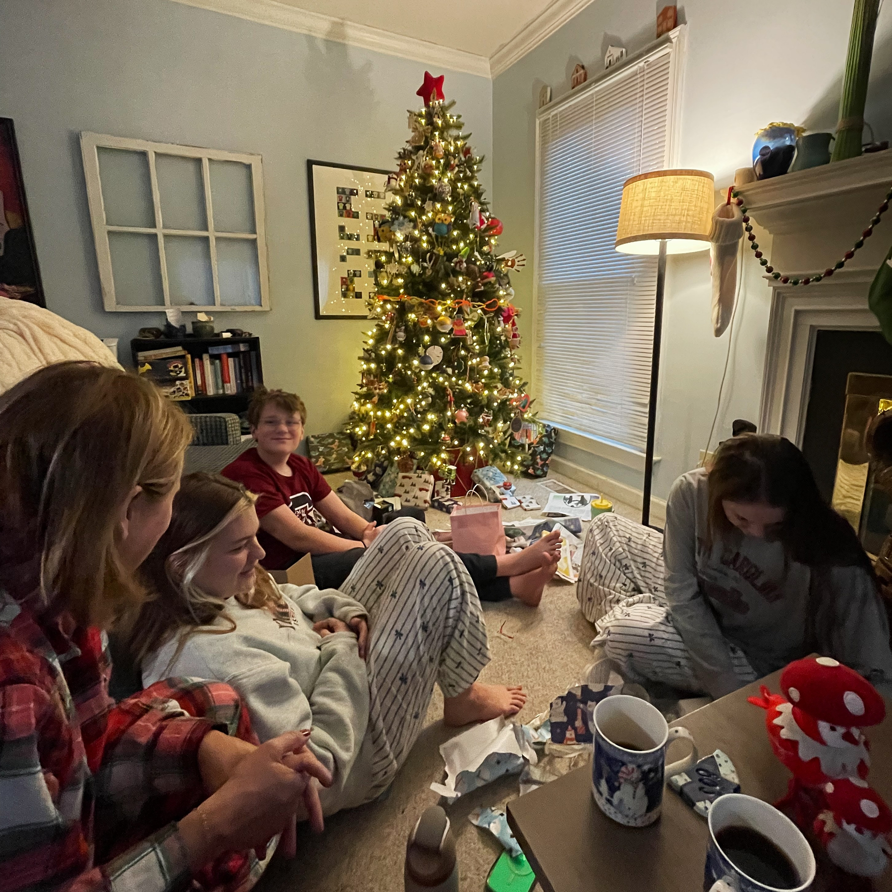 A family in pajamas are sitting around a Christmas tree with presents, in a cozy room decorated for the holidays.