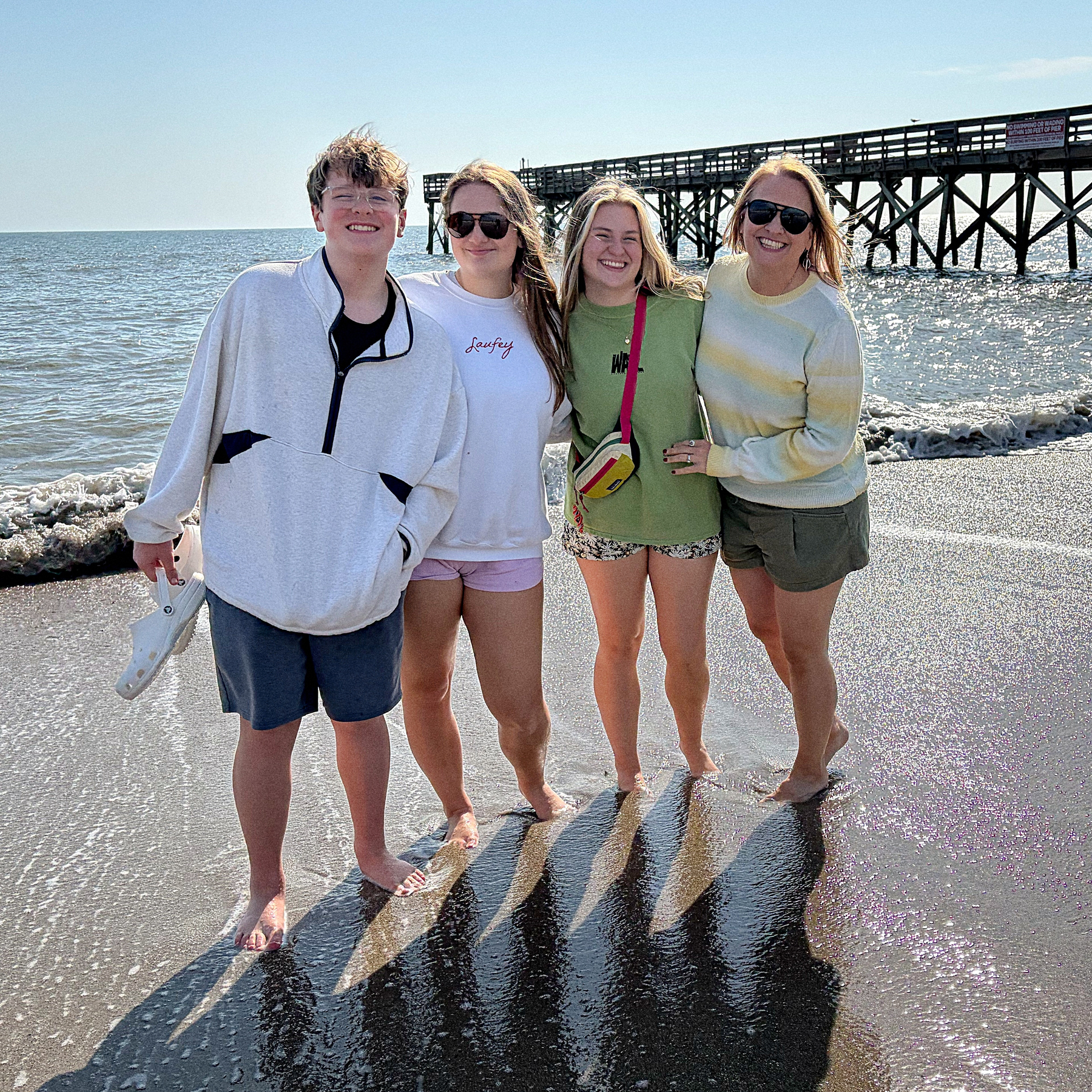 Four people are standing on a beach near the water with a pier in the background.
