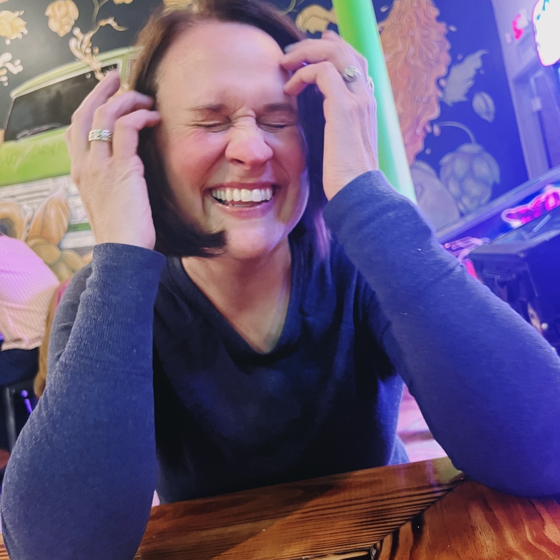 A person sits at a wooden table, laughing with eyes closed and hands on their head, in a colorful indoor setting.