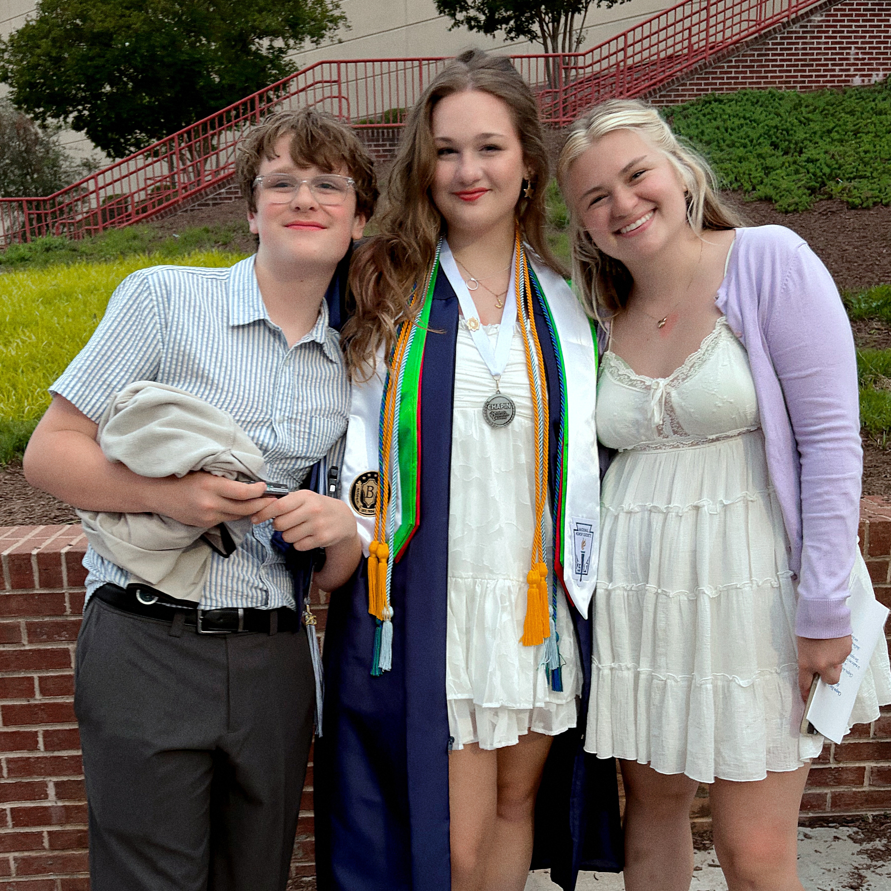 Three people are posing together, one in graduation attire, all smiling outdoors near a building and steps.