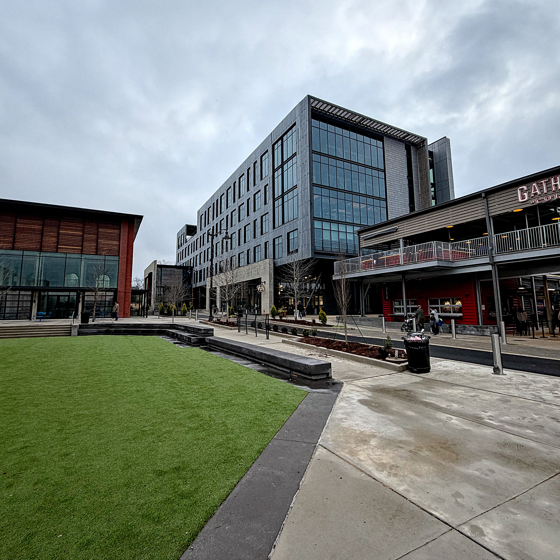 A modern urban plaza features a large building with glass windows, a grassy area, and a restaurant with outdoor seating.