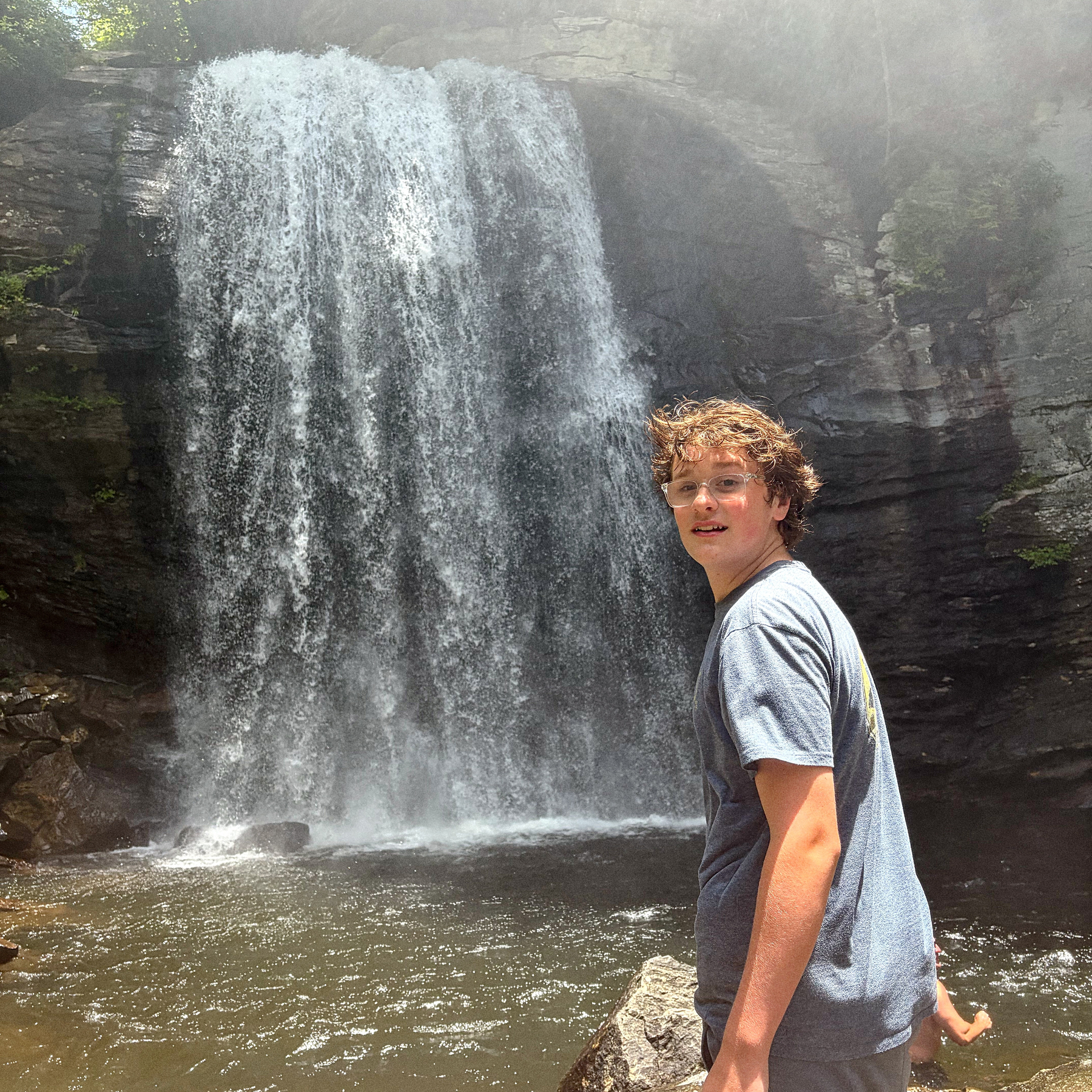 A young person wearing glasses and a gray t-shirt is standing on a rock in front of a large waterfall cascading down a rocky cliff.