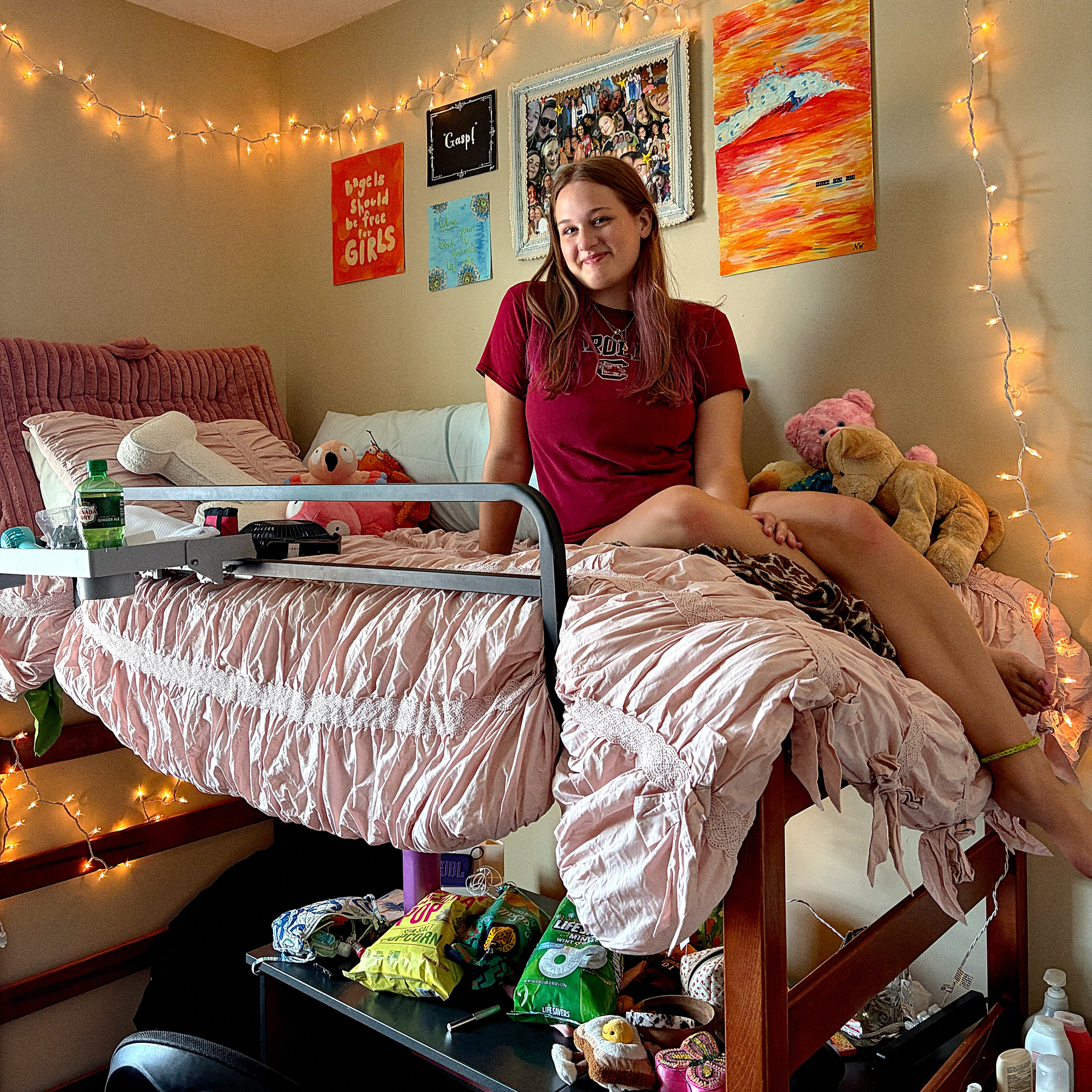 A young woman is sitting on a bed in a cozy, decorated bedroom with string lights, posters on the wall, stuffed animals, and various snacks on a table underneath.