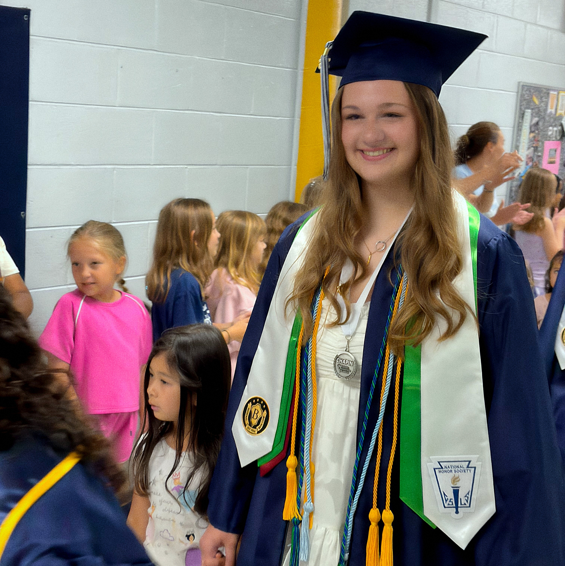 A graduate in a cap and gown stands smiling in a hallway while young children line up in the background.