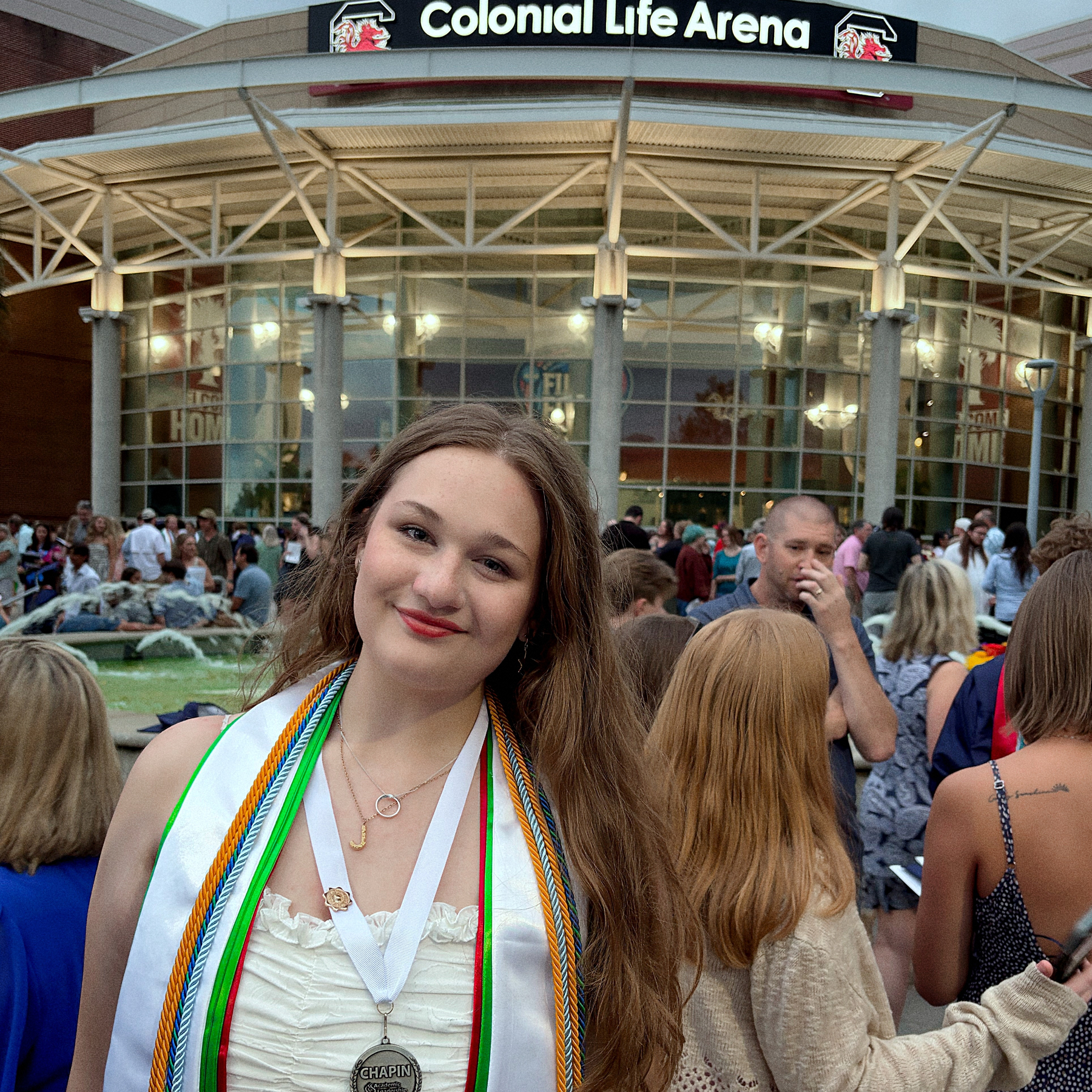 A person wearing a graduation gown and medals smiles in front of a crowd at an event outside Colonial Life Arena.