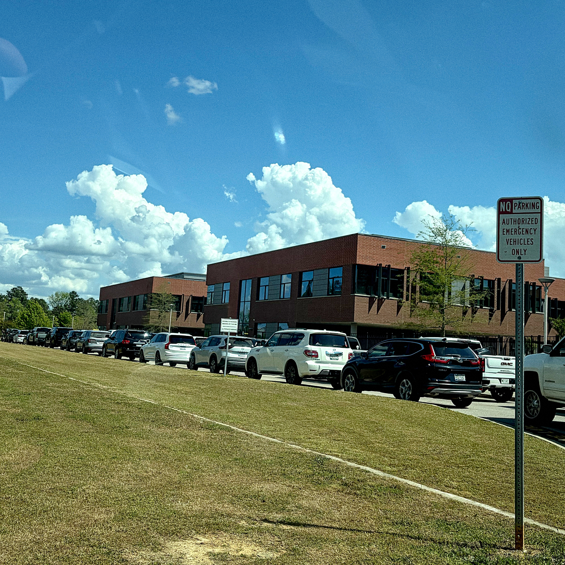 A line of parked cars is visible along a grassy area near a brick building under a blue sky with fluffy clouds.