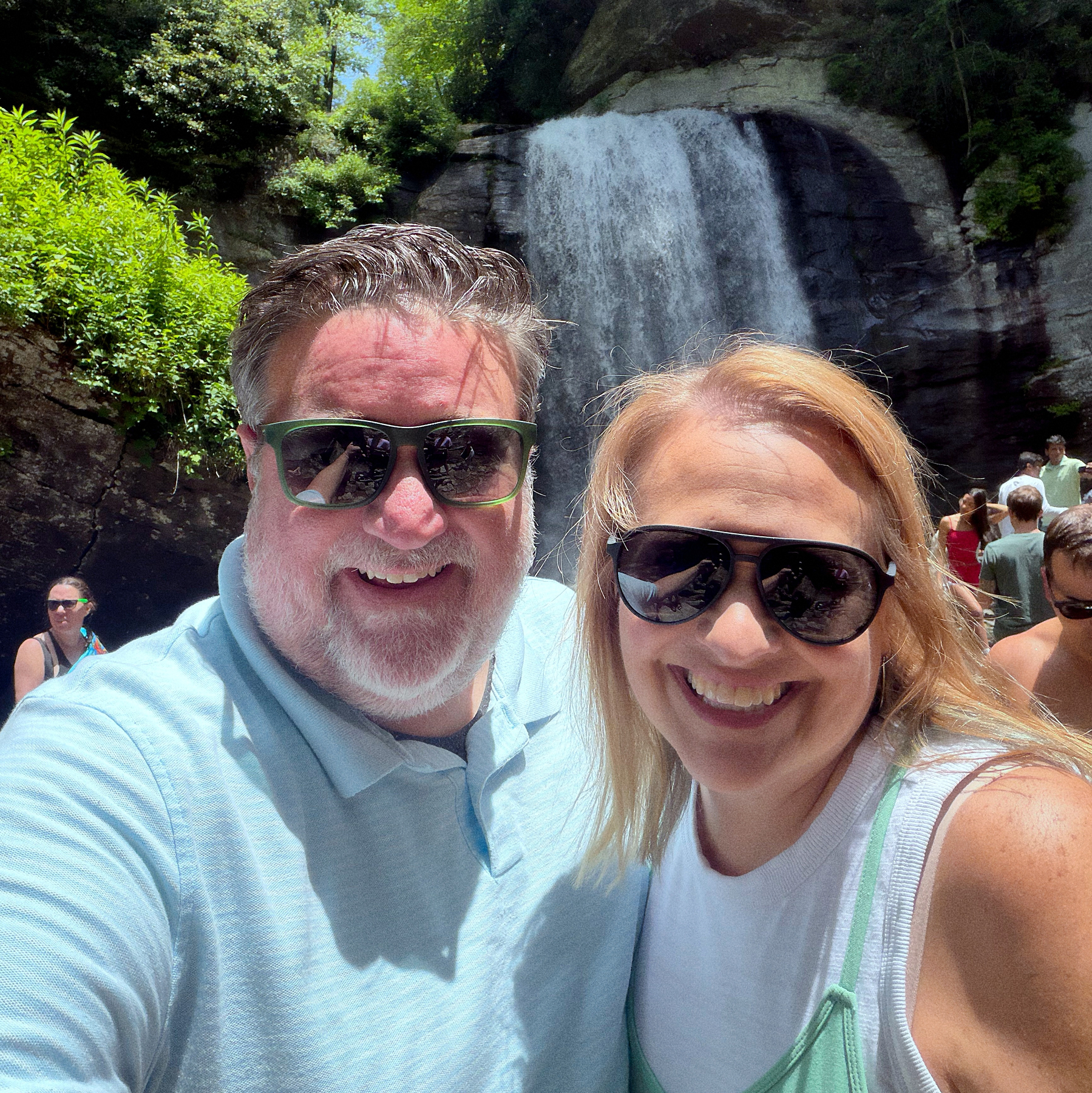 A man and a woman wearing sunglasses smile together in front of a waterfall surrounded by lush greenery.