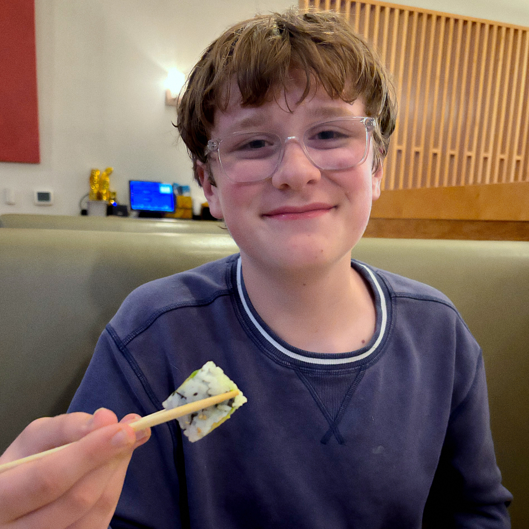 A smiling person with glasses holds a piece of sushi using chopsticks while seated in a restaurant.