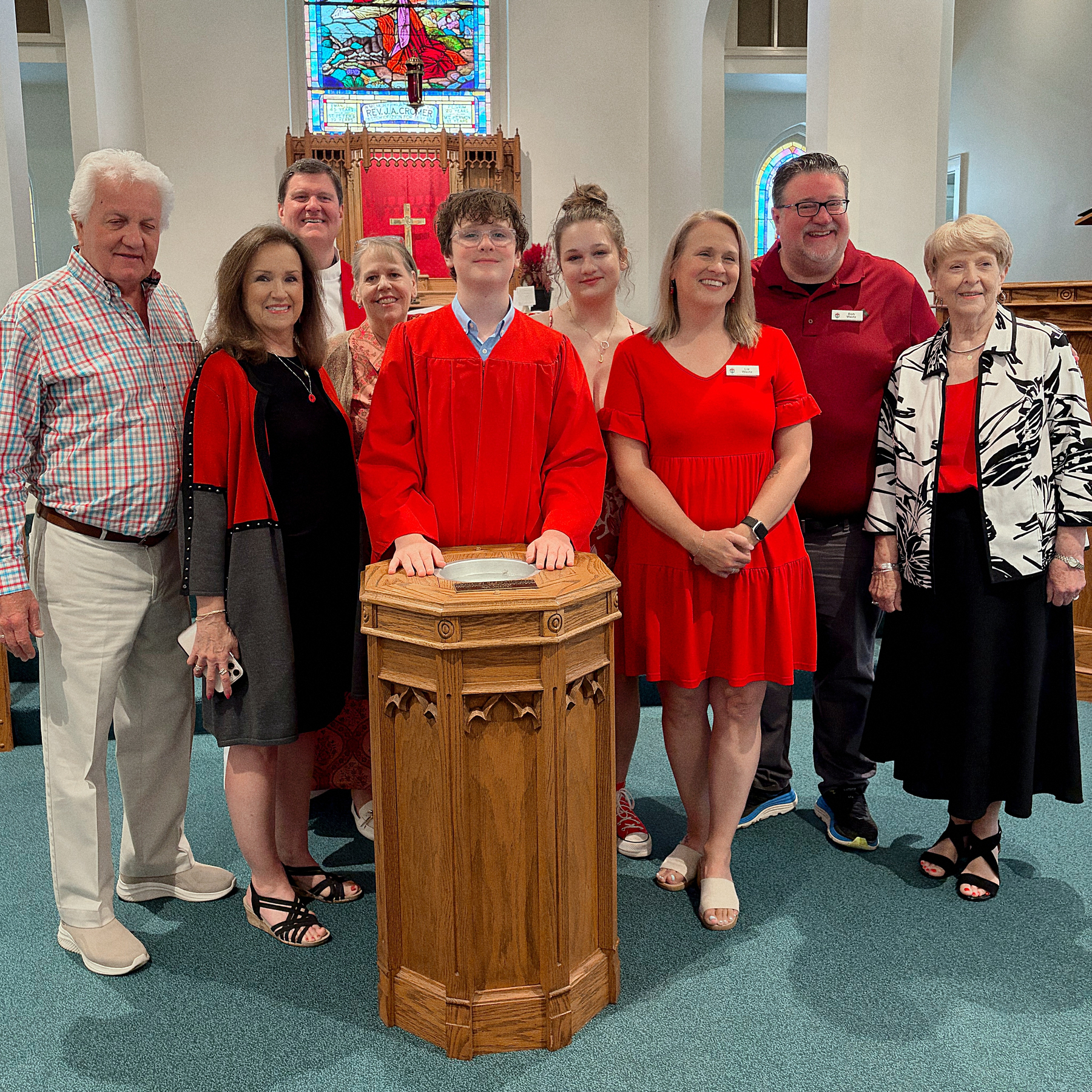 A group of people, wearing a mix of black, red, and white clothing, stand together smiling in a church setting with a young man in a red gown at the center.