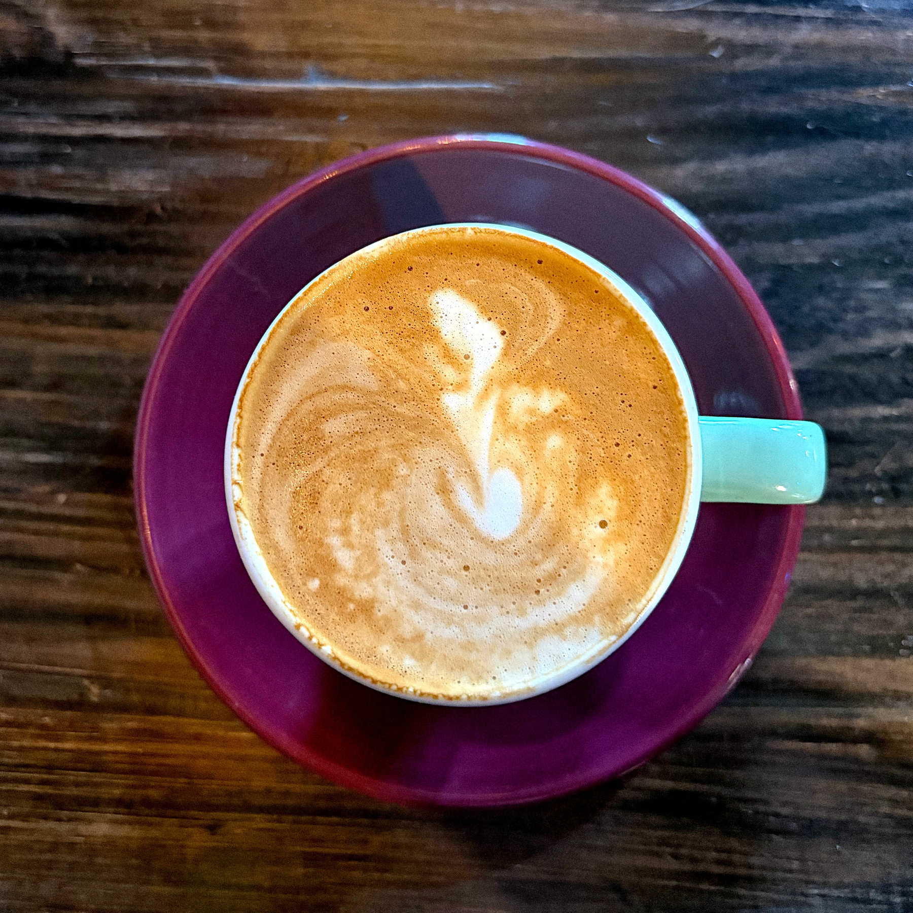 A cortado with heart-shaped latte art sits on a purple saucer atop a wooden surface.