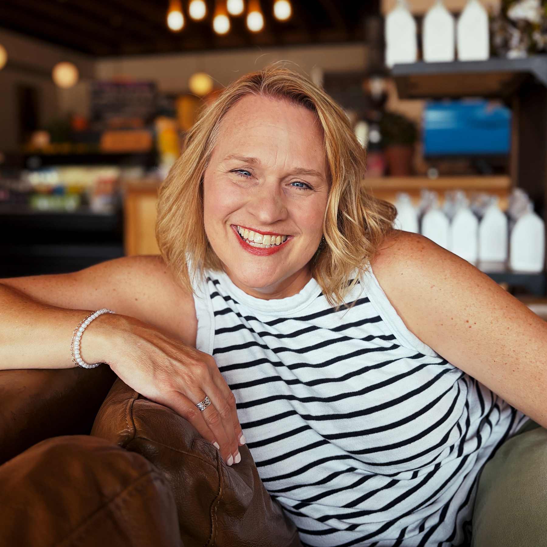 A smiling woman with blonde hair is leaning on a brown couch in a warmly lit, cozy café environment.