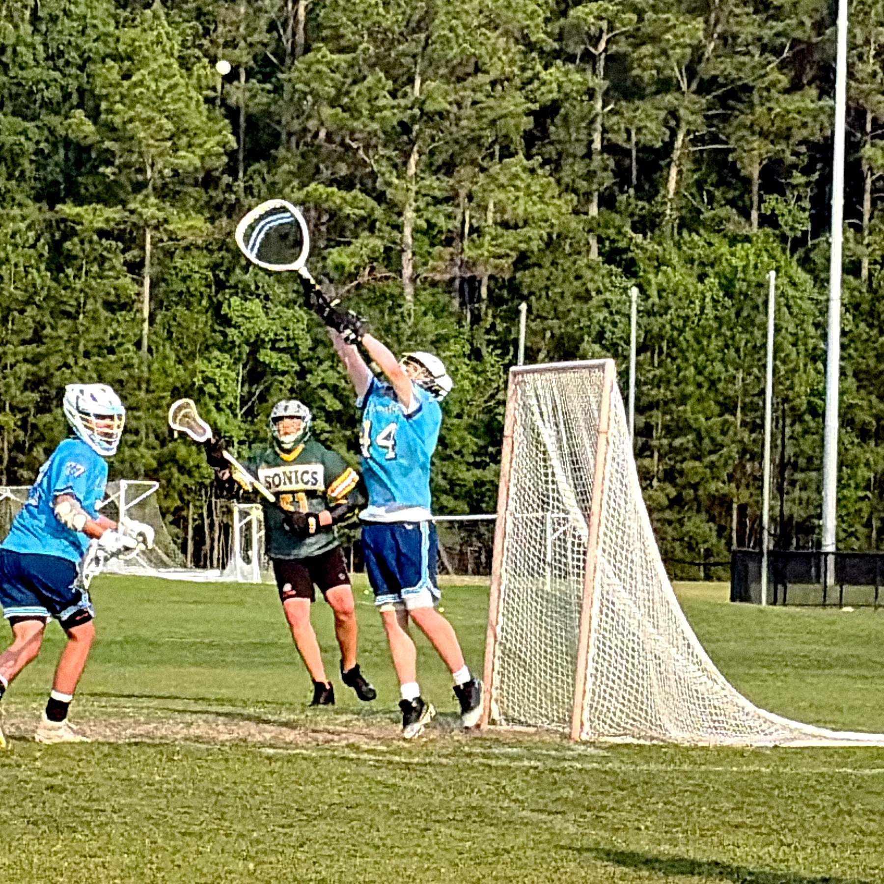 A lacrosse player in blue attempts to catch the ball near the goal while two other players and a goalkeeper look on in a grassy field.