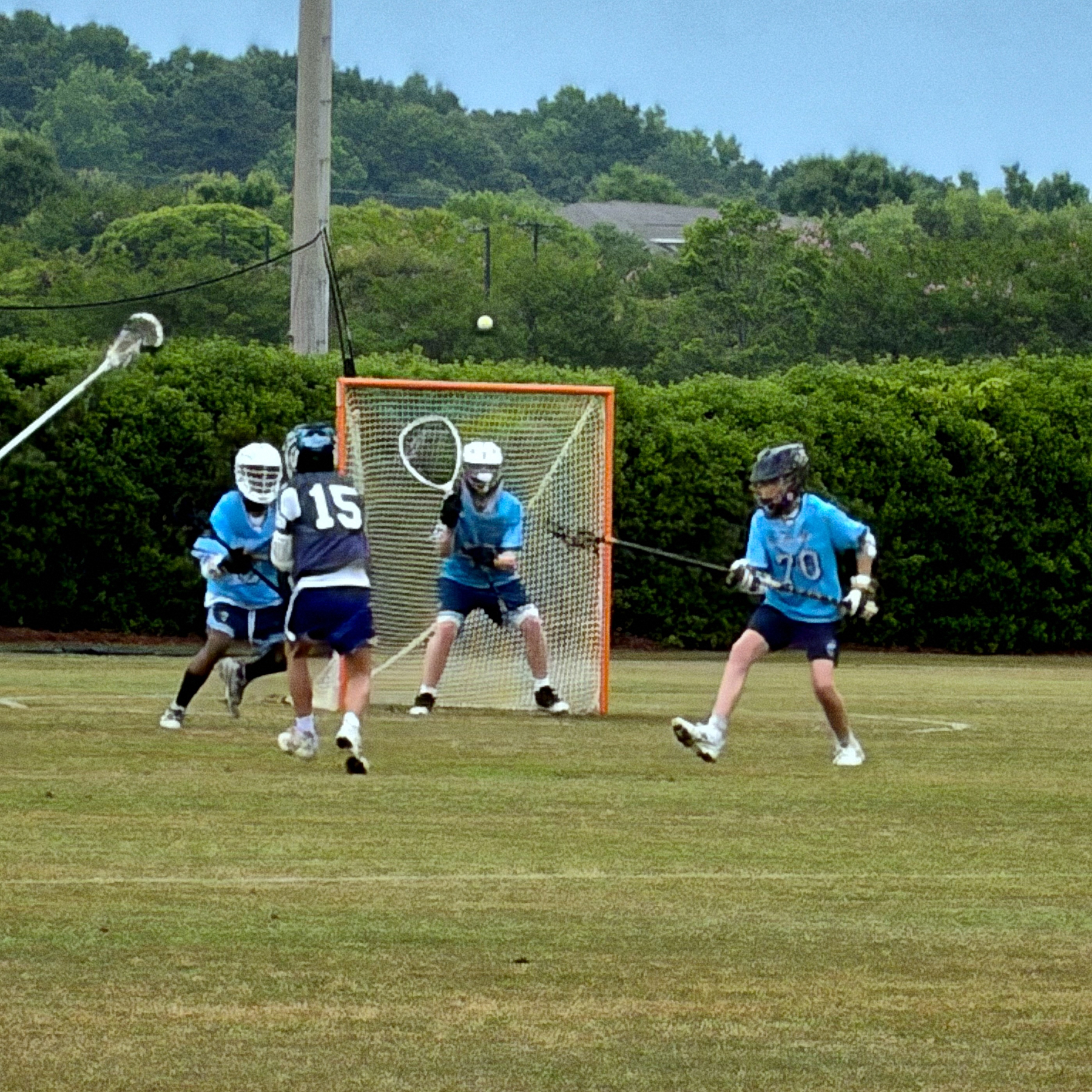 Four young lacrosse players, wearing blue and white uniforms and protective gear, engage in a game on a field with a net behind them.
