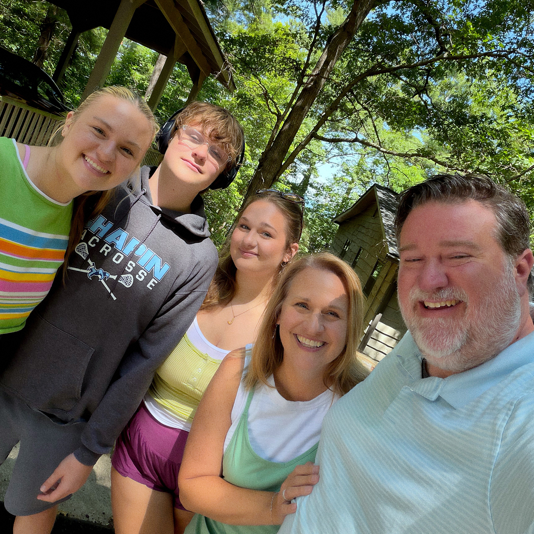 A group of five people, consisting of two men and three women, are smiling and posing together outside in a sunny, wooded area near some buildings.