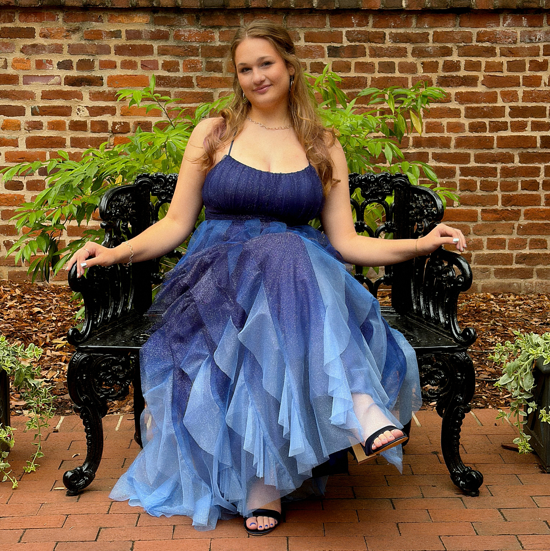 A woman wearing a flowing blue dress sits on a black ornate bench against a brick wall backdrop.