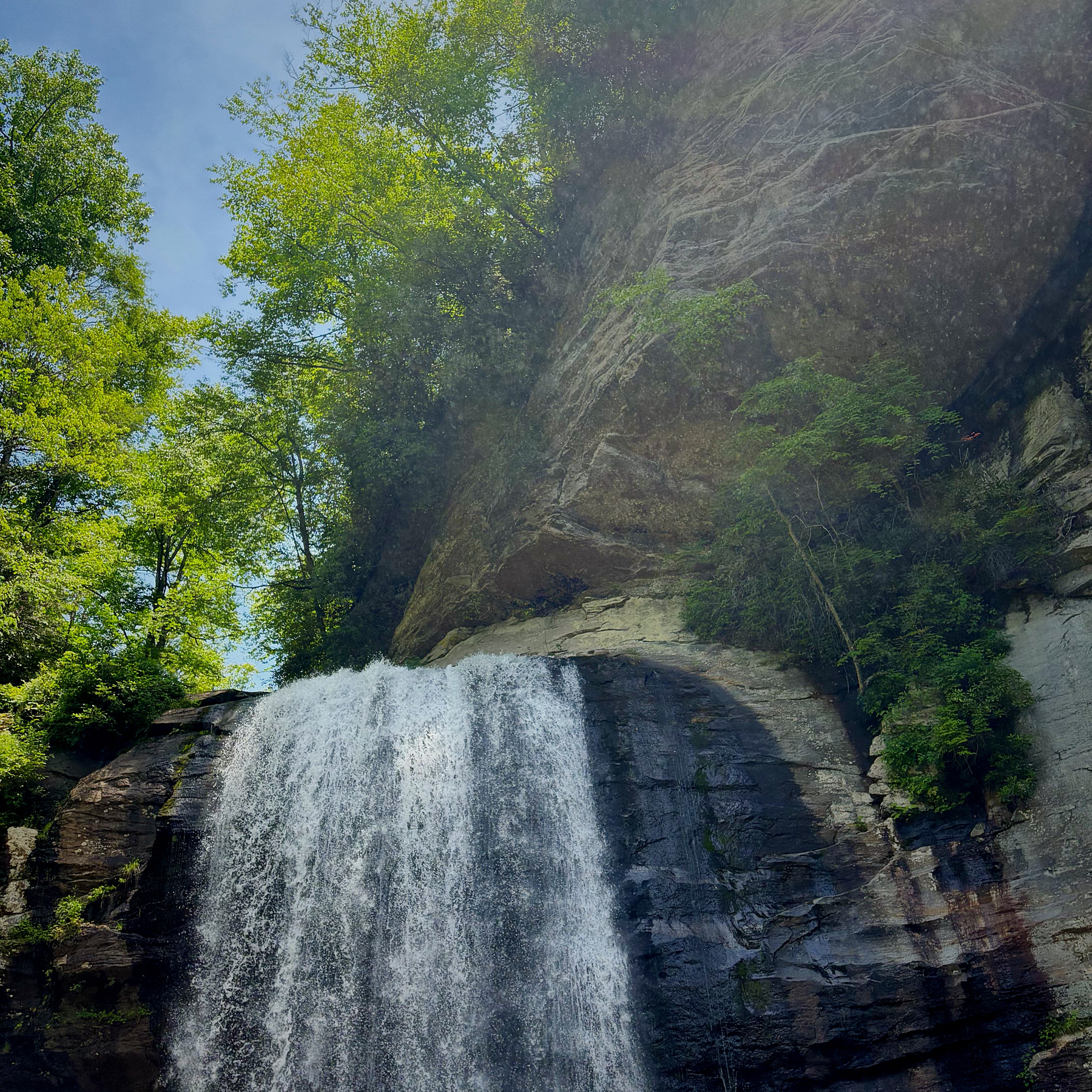 A waterfall cascades down a rocky cliff surrounded by lush green trees under a clear blue sky.