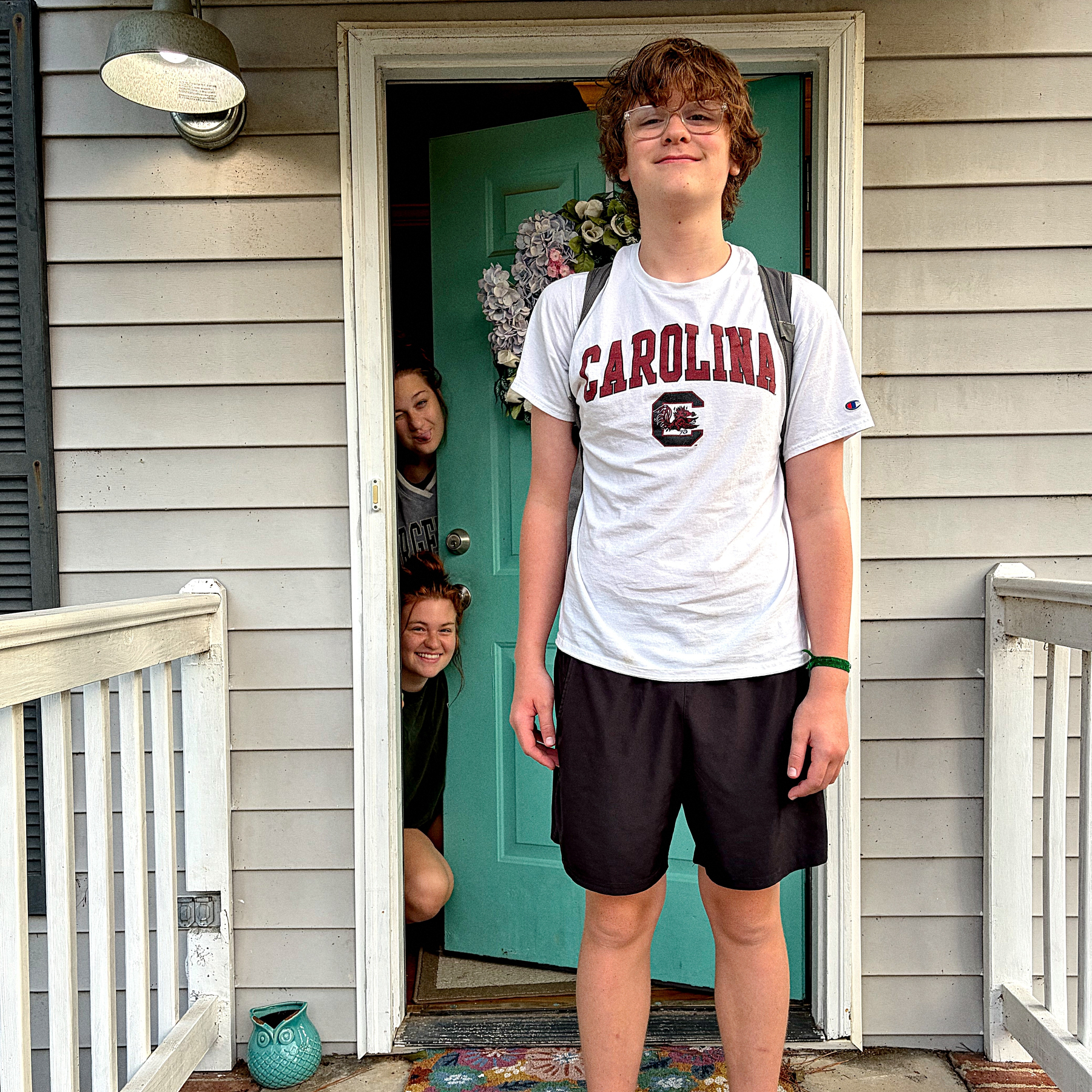 A boy wearing a CAROLINA t-shirt, glasses, and a backpack stands smiling on a porch while two people peek out from behind a partially open door.