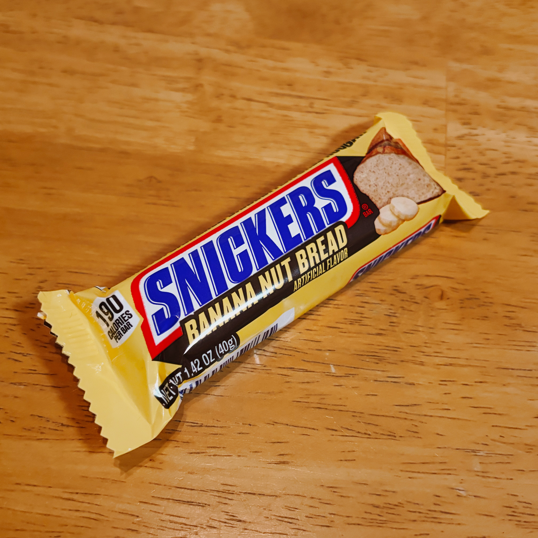 A Snickers Banana Nut Bread flavored candy bar with 190 calories is placed on a wooden surface.