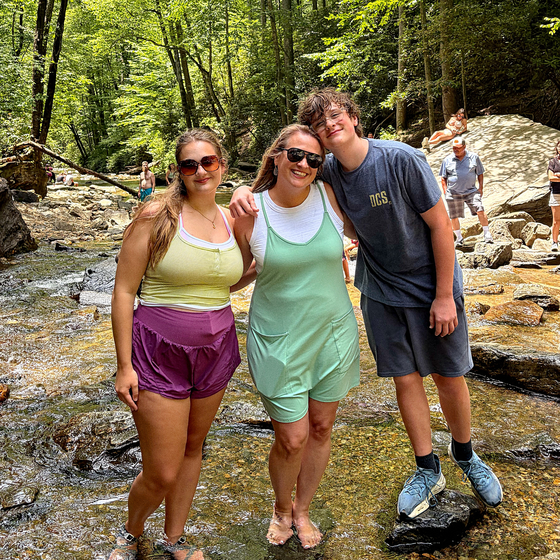 Three people posing and smiling together while standing in a shallow rocky stream surrounded by lush green forest.