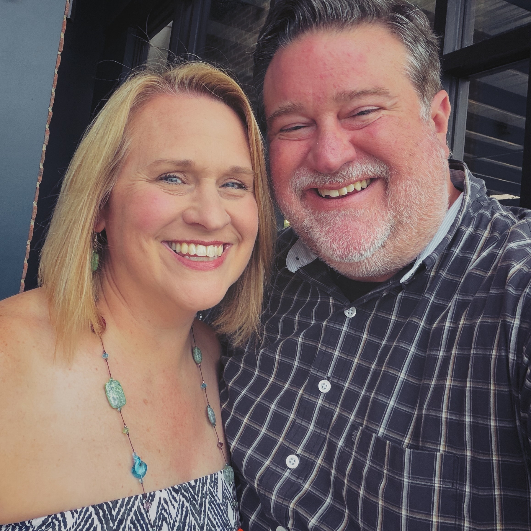 A smiling couple poses closely together, with the woman wearing a sleeveless patterned top and the man in a checkered shirt.