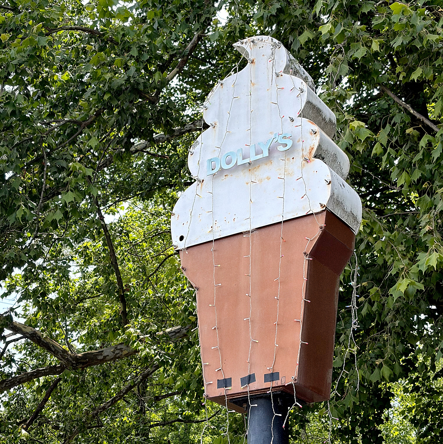 A large ice cream cone sign with the word DOLLY'S on it is mounted on a pole surrounded by leafy green trees.