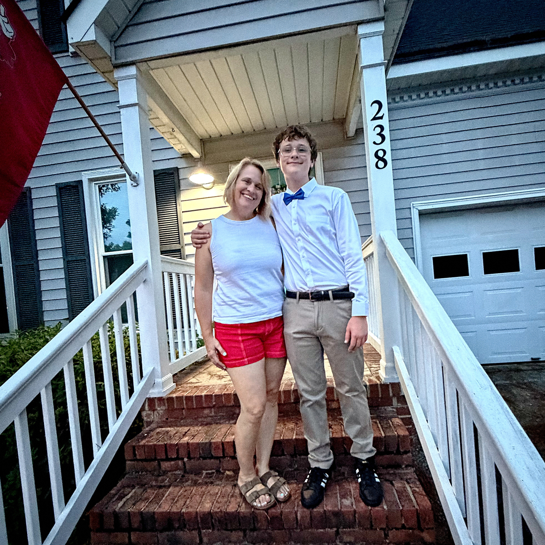 A smiling woman in a white sleeveless top and red shorts stands with a young man dressed in a white shirt, blue bow tie, and beige pants on the brick stairs in front of a house.