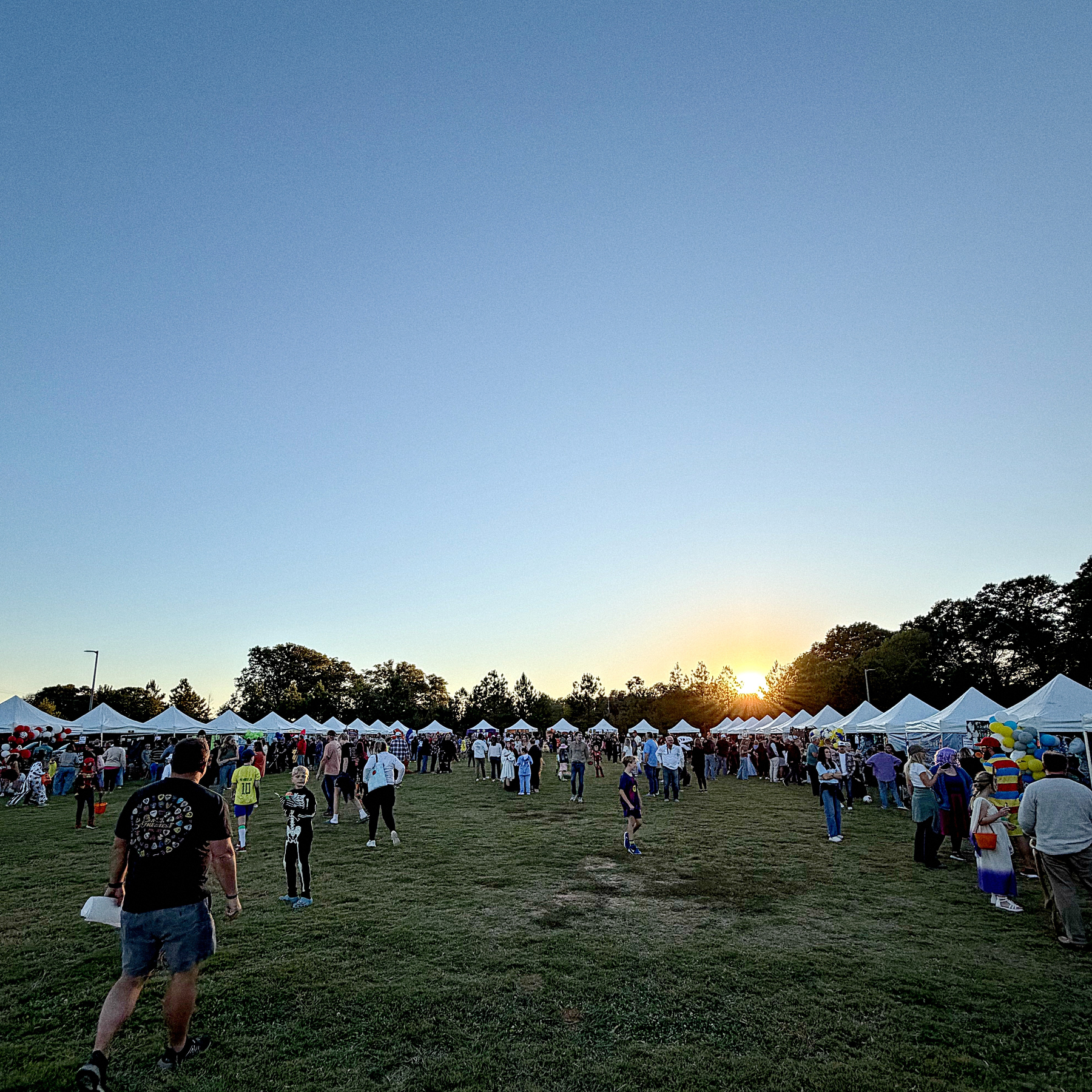 A crowd of people is walking and mingling among white tents set up on a grassy field at sunset.