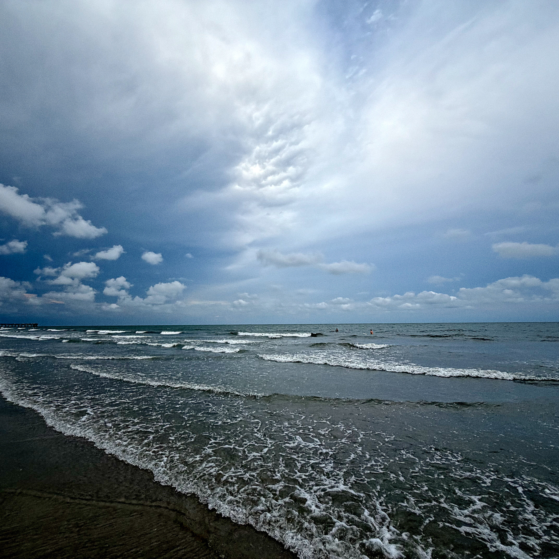 A cloudy sky is reflected over gentle waves rolling onto a sandy beach.