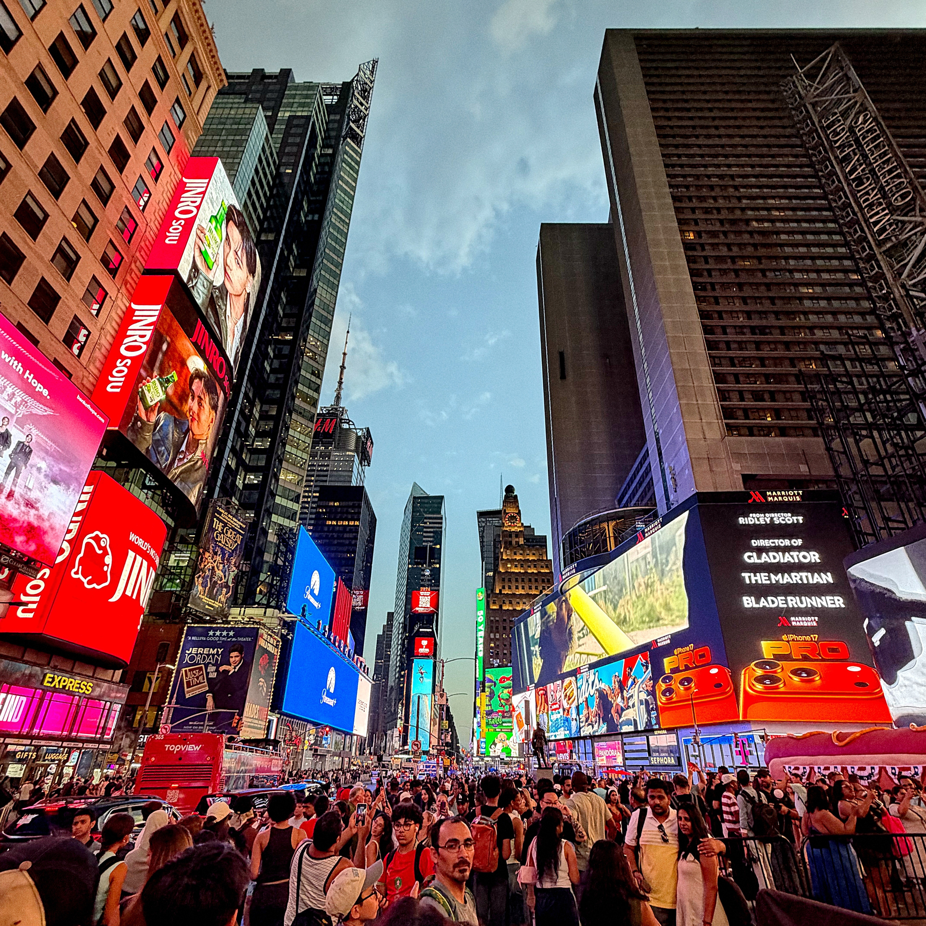A bustling crowd fills a city square surrounded by tall buildings adorned with numerous colorful electronic billboards and advertisements against a partly cloudy sky.