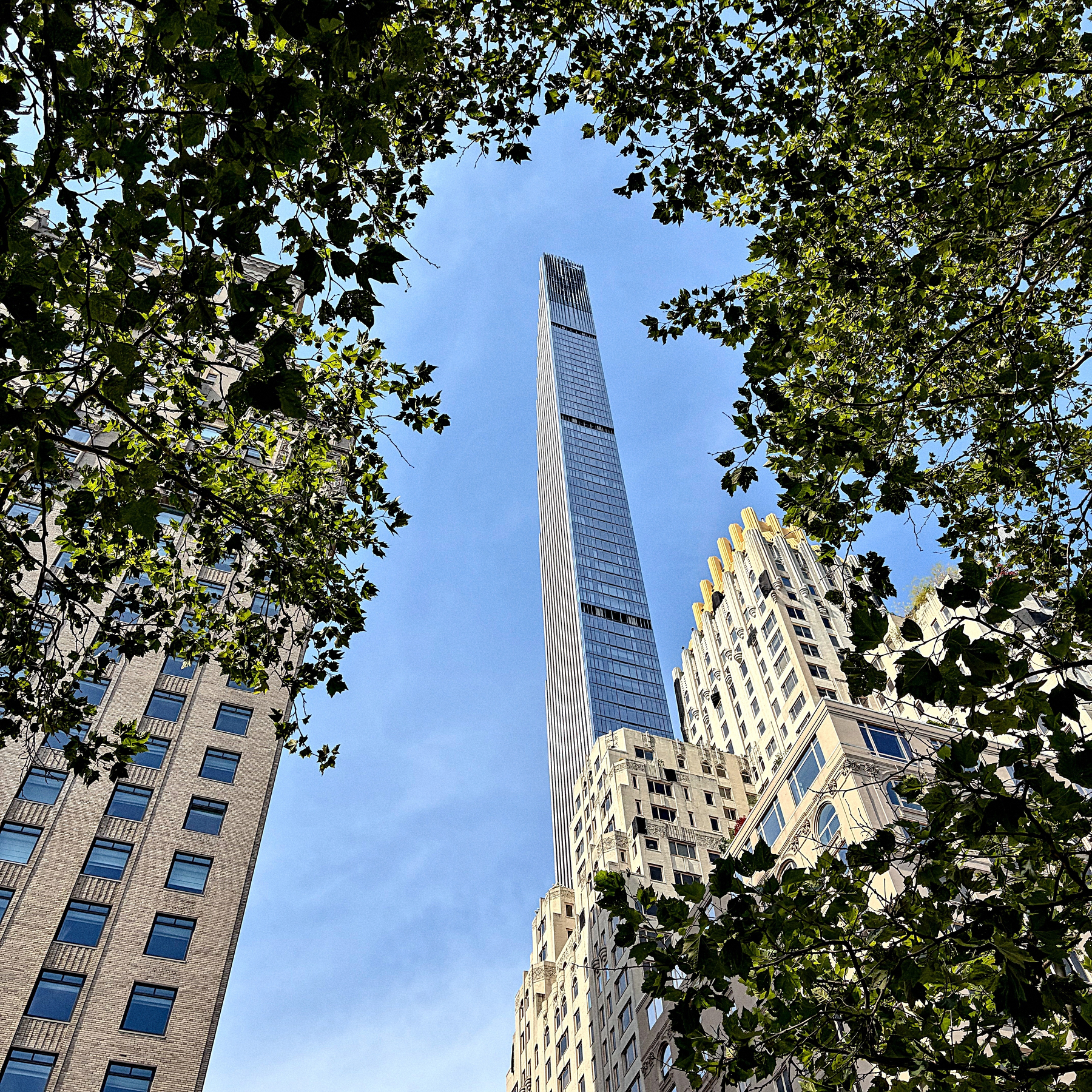 A tall, slender skyscraper is framed by surrounding buildings and tree branches against a clear blue sky.