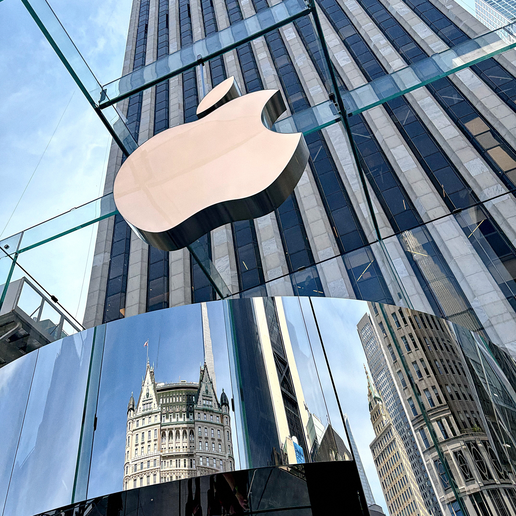 A large Apple logo is displayed on a glass structure with a reflection of surrounding tall buildings in the shiny surface below.