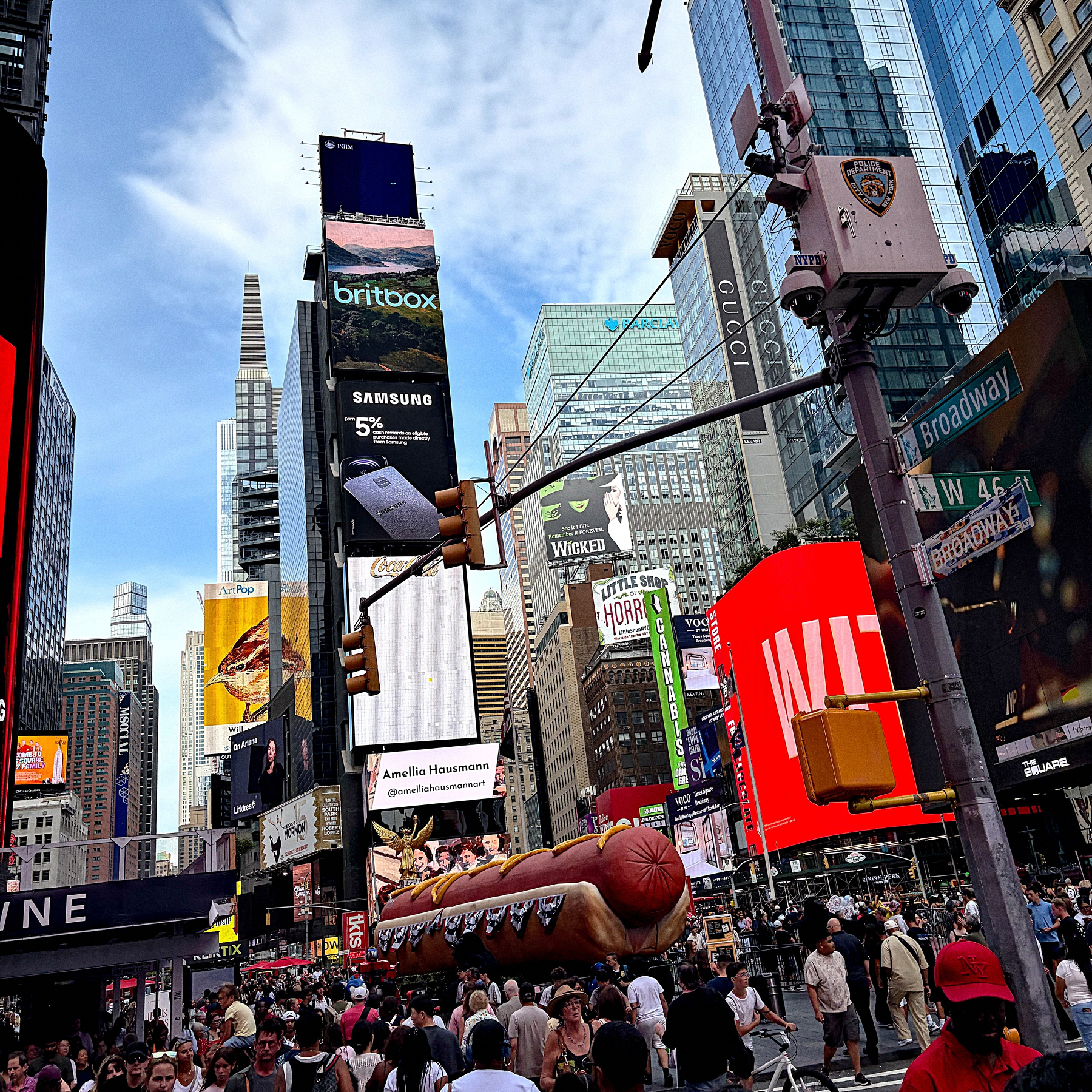 A crowded street scene in Times Square, New York, features vibrant billboards, skyscrapers, and a large inflatable hot dog among the bustling crowd.